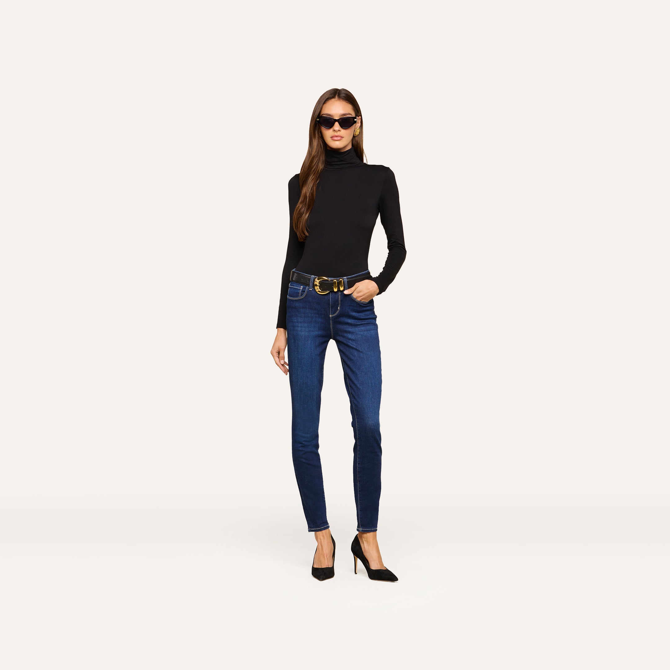 A woman wearing sunglasses, a black long-sleeve turtleneck, a belt, and black high heels stands against a plain light background. She completes her outfit with the Marguerite Skinny Jean—blue high rise skinny jeans crafted from stretch denim.