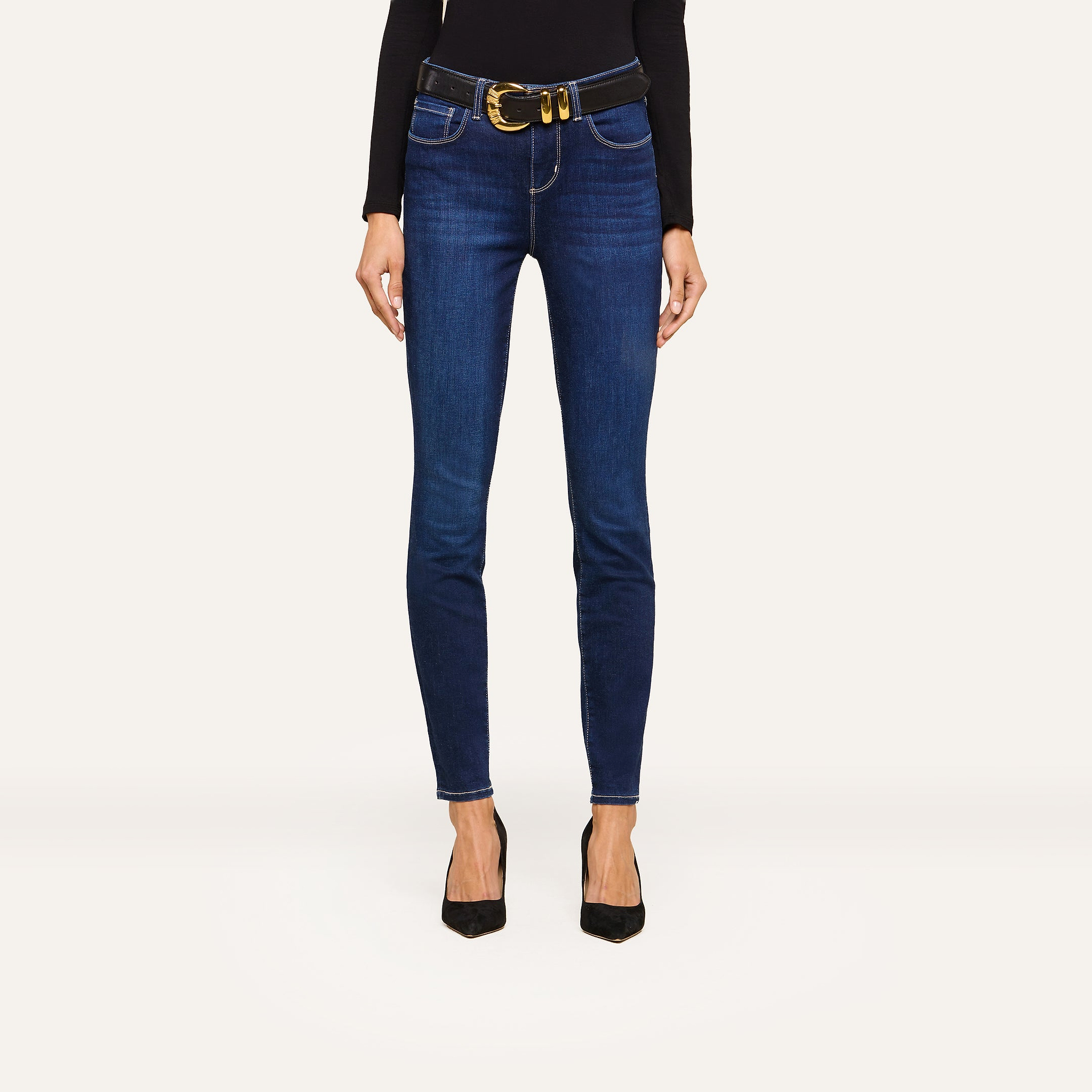 Person wearing dark blue skinny jeans, a black long-sleeve top, a wide black belt with a large gold buckle, and sleek black pointed-toe heels, standing against a plain white background.