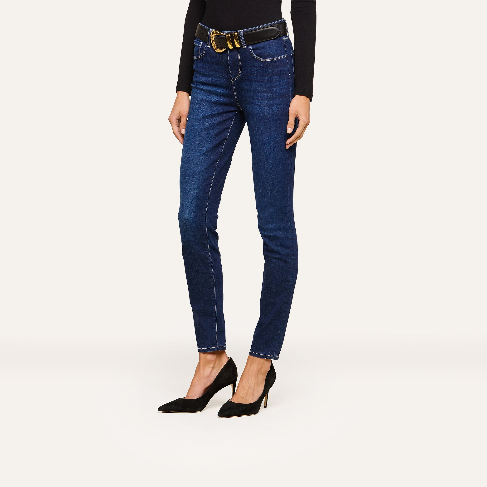 Person wearing dark blue skinny jeans with a black belt, paired with a fitted black long-sleeve top and black high-heeled shoes, standing against a plain white background. Face is not visible.