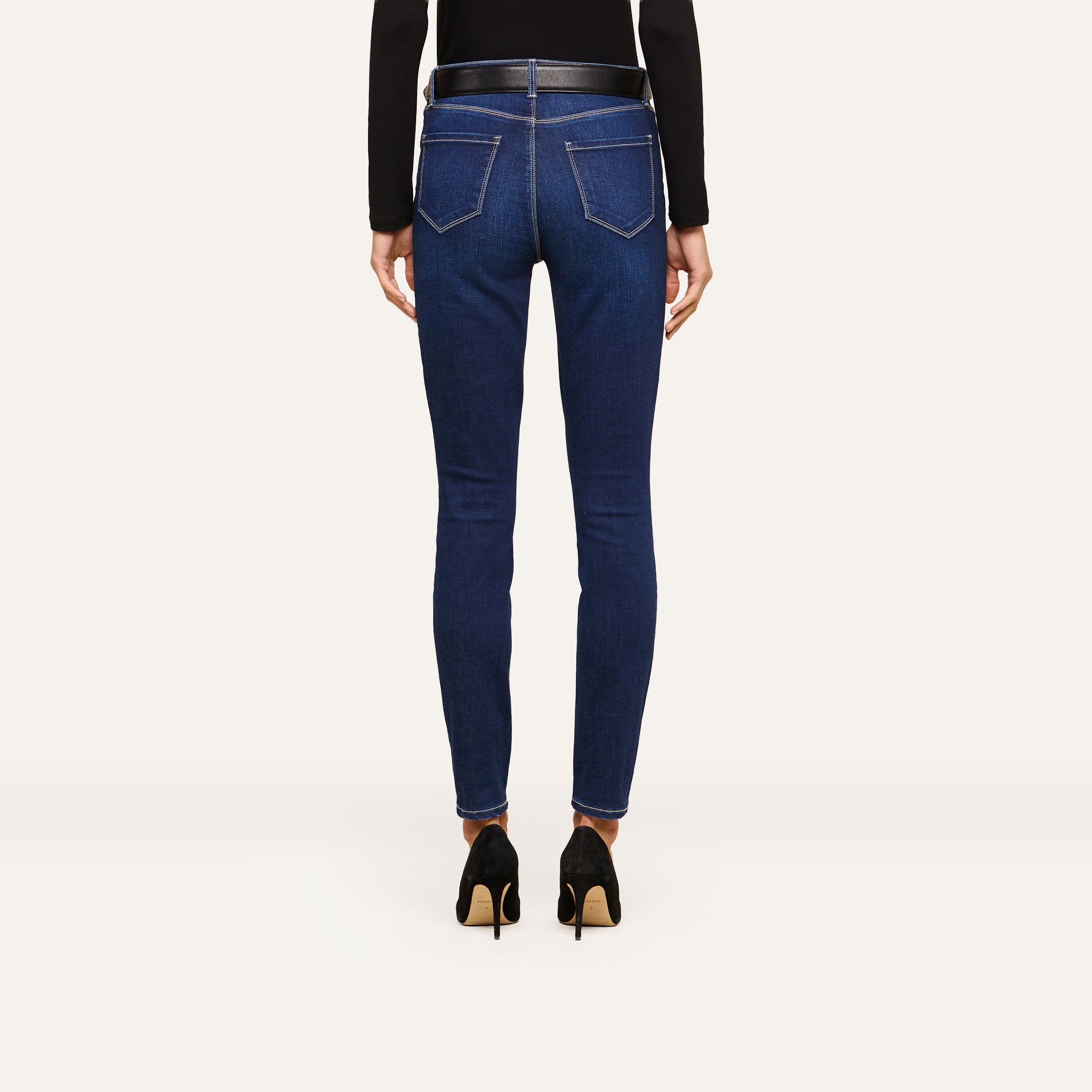 A person wearing a black long-sleeve top, high-waisted blue skinny jeans, and black high heels is shown from the back, standing against a plain white background. The sleek outfit highlights modern fashion trends.