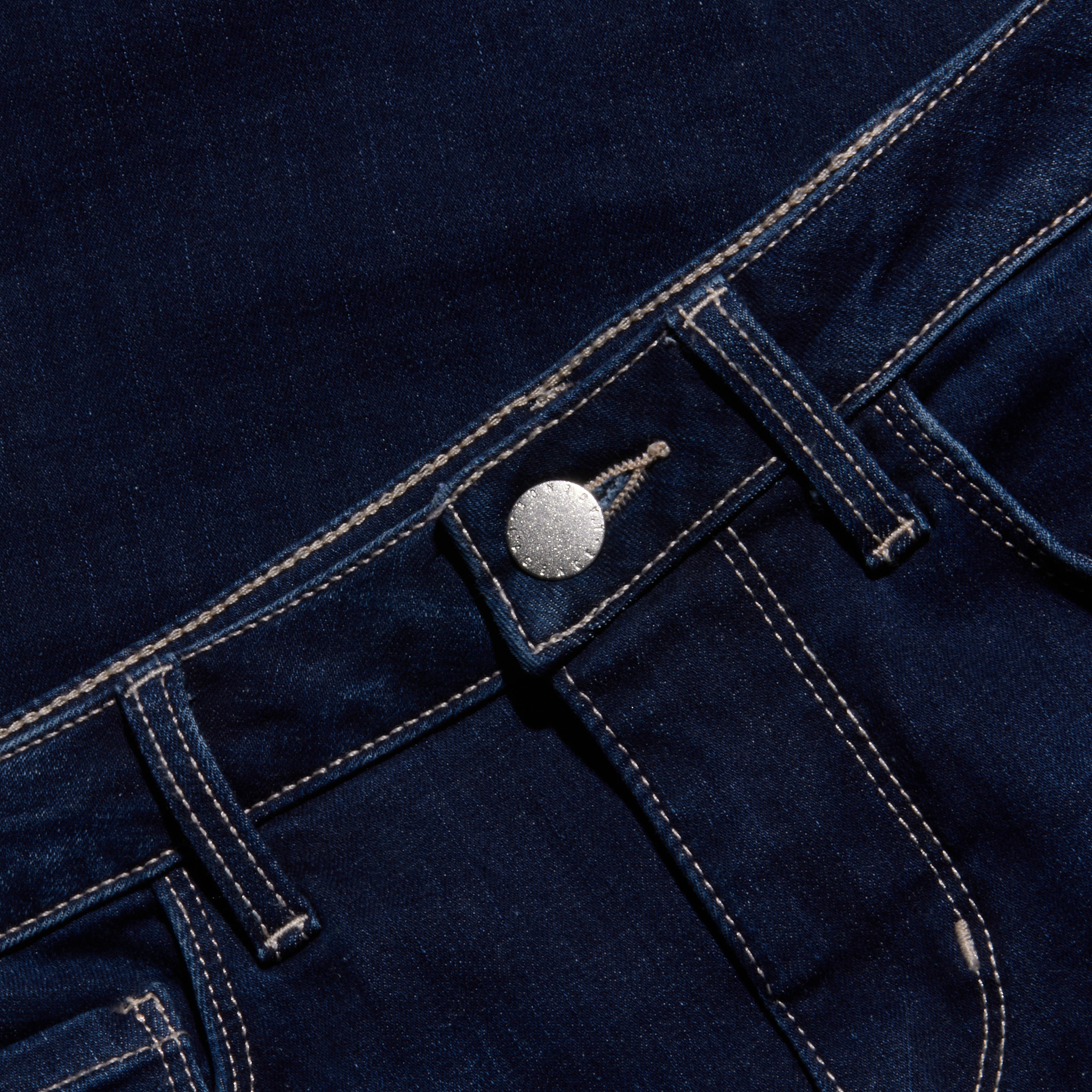 Close-up of the waistband and front pocket of dark blue denim jeans, featuring a silver button, classic belt loops, and visible white stitching for a timeless look.