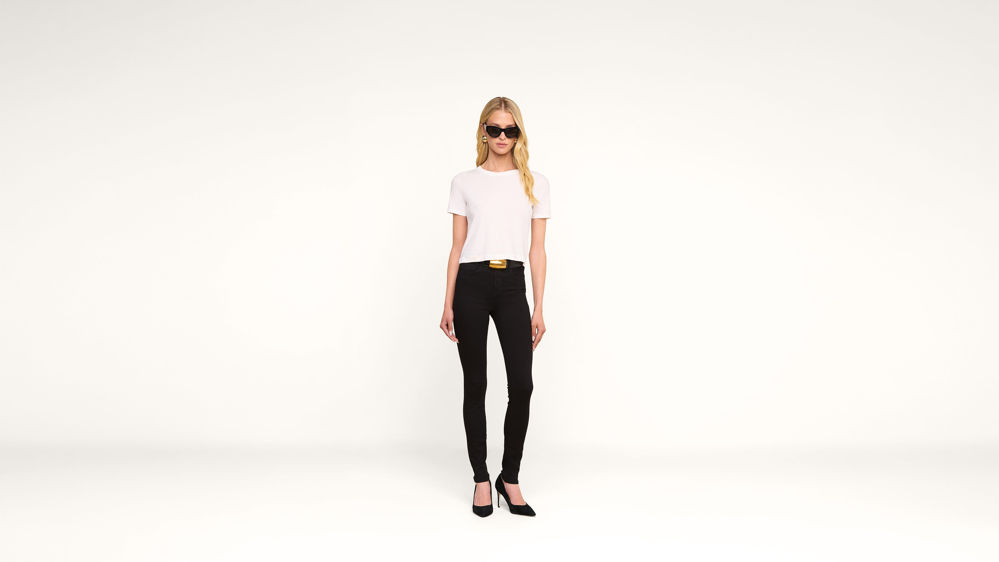 A woman wearing sunglasses, a white t-shirt, black pants, and black heels stands against a plain white background.
