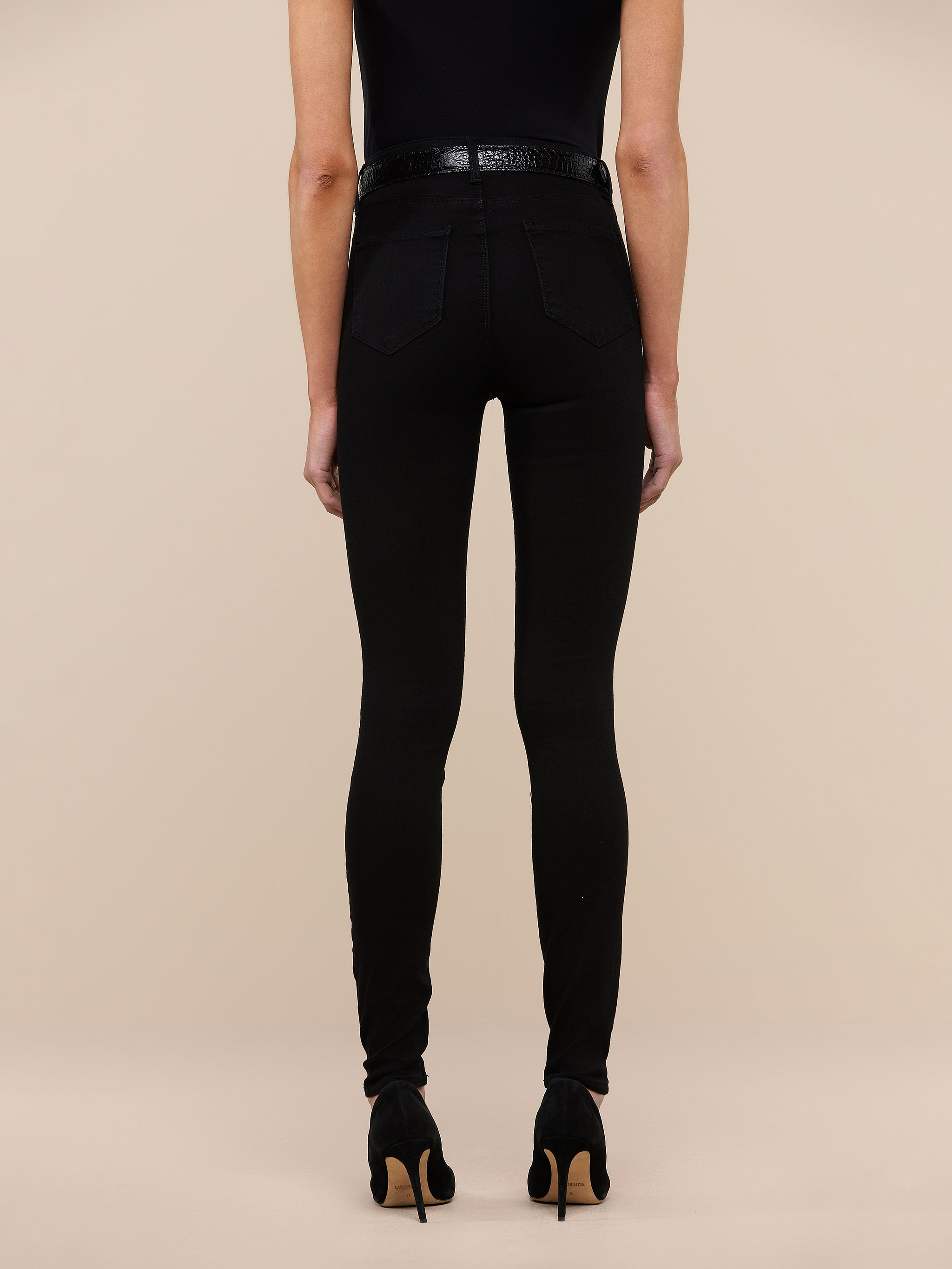 A person stands with their back to the camera against a plain white background, wearing the Marguerite Skinny Jean in black high-recovery fabric, styled with black high heels and a white top.