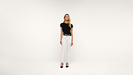 A woman stands facing forward against a plain white background, wearing a black t-shirt, white pants, a black belt with a gold buckle, and black heels.
