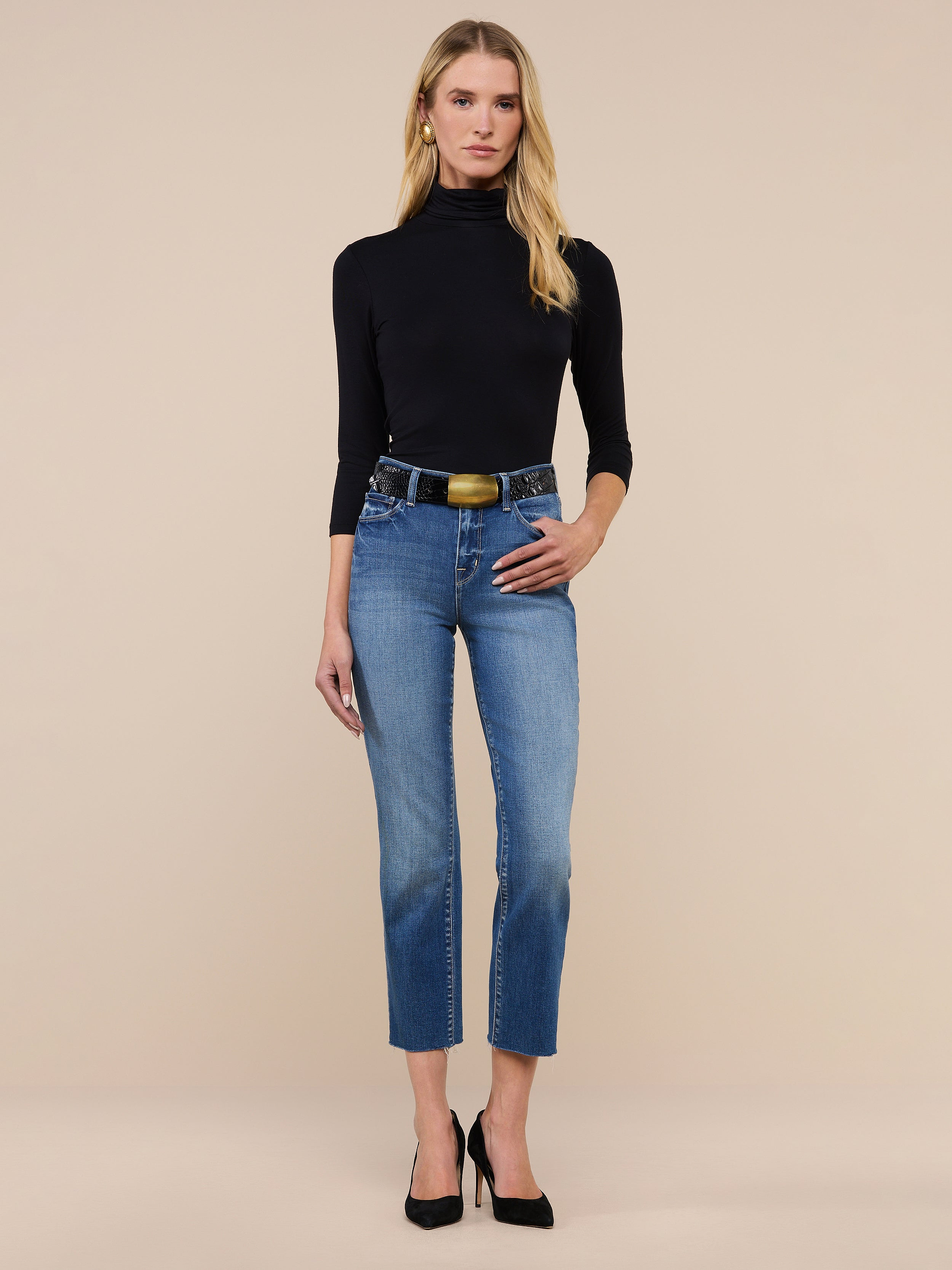 A woman stands against a plain background wearing a black turtleneck, black heels, and the Sada Slim-Leg Cropped Jean—a pair of blue high-rise cropped jeans crafted from premium stretch denim with a black belt. She has long blonde hair and her left hand rests in her pocket.