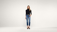 A woman stands against a plain white background, wearing a black long-sleeve turtleneck, blue jeans, a black belt with a gold buckle, and black heels. She has blonde hair and a neutral expression.
