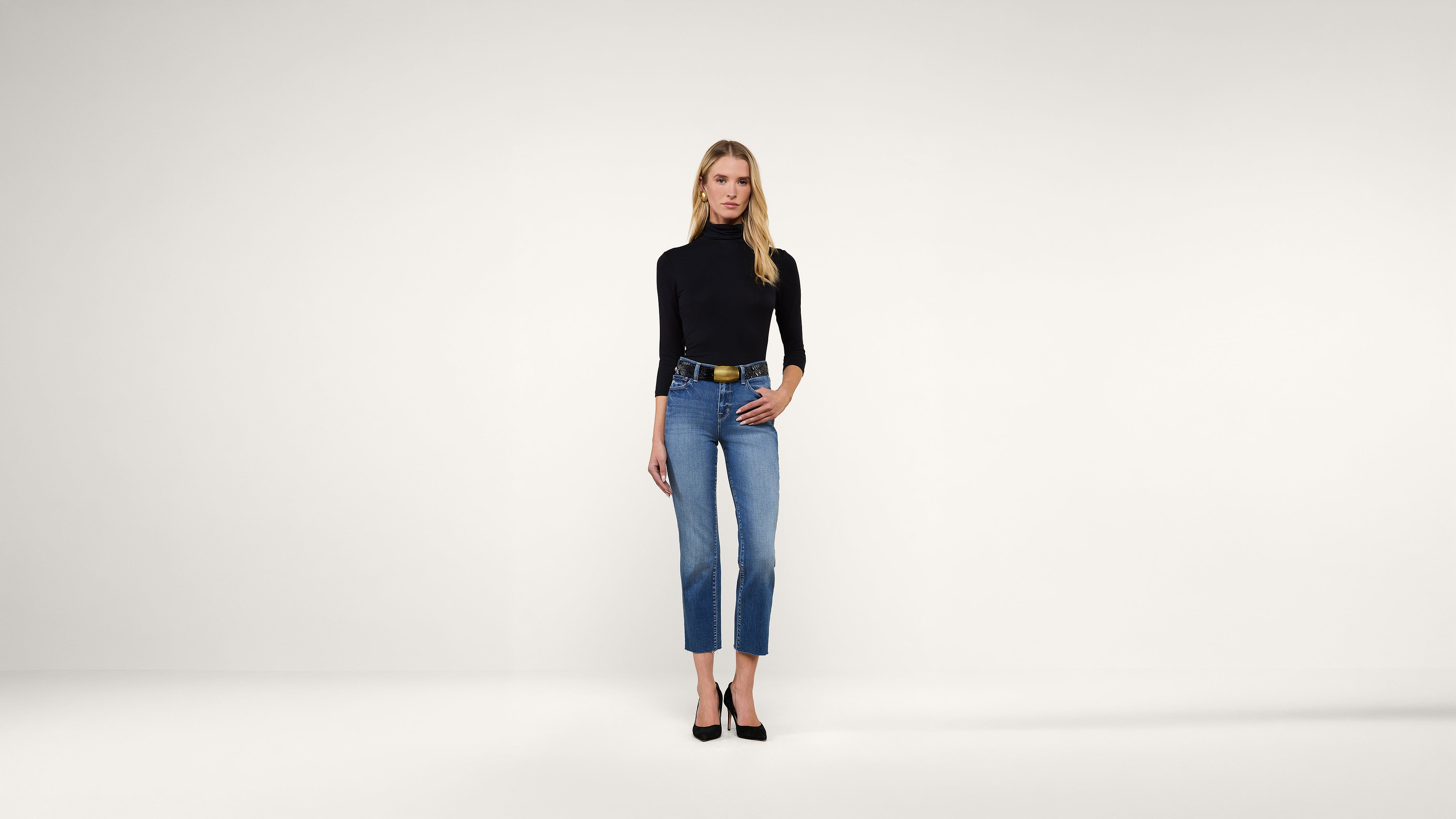 A woman stands against a plain white background, wearing a black long-sleeve turtleneck, blue jeans, a black belt with a gold buckle, and black heels. She has blonde hair and a neutral expression.