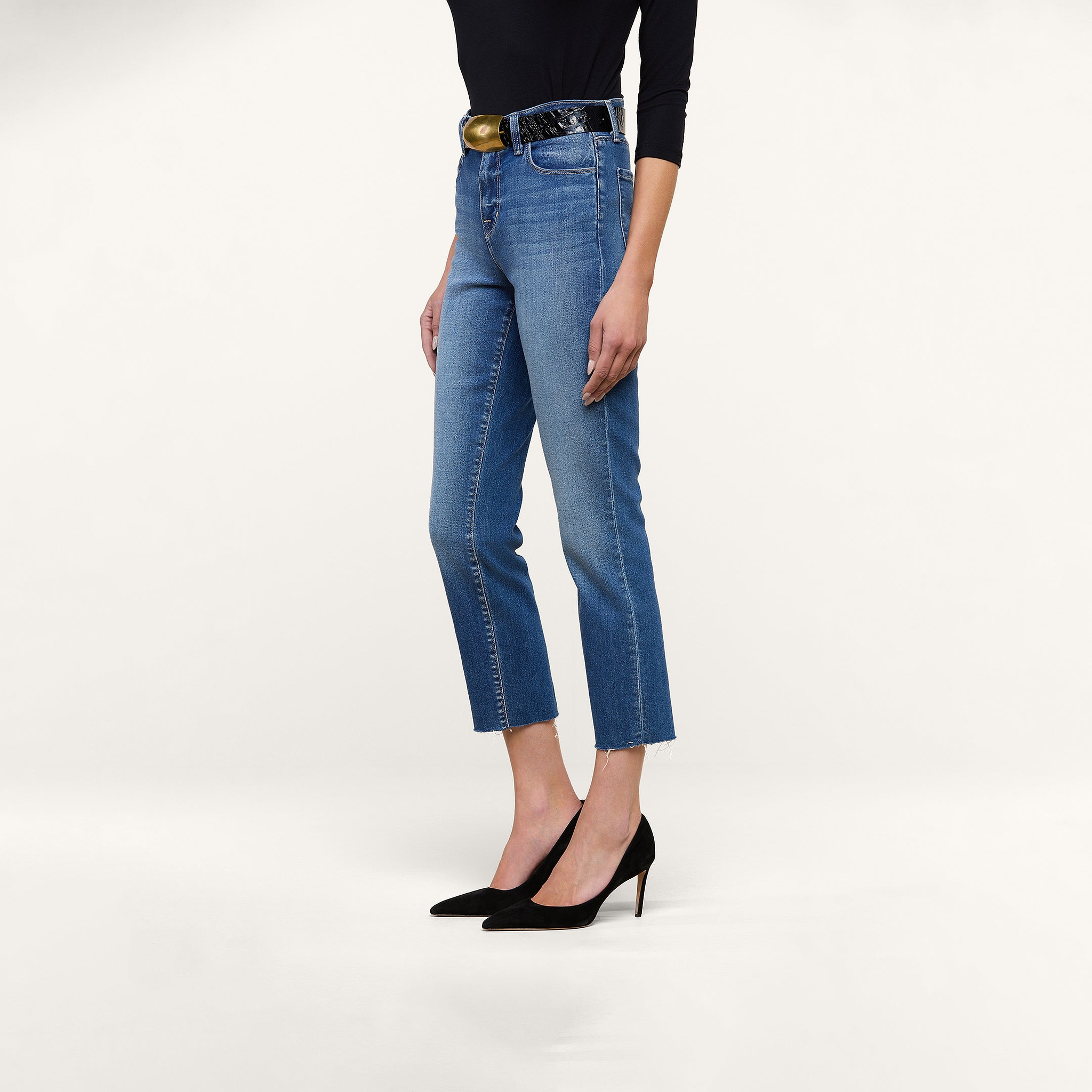 A person wearing the Sada Slim-Leg Cropped Jean in blue—featuring a high-rise, slim straight leg, and premium stretch denim—paired with a black long-sleeve top, black belt, and black high-heeled shoes stands against a plain white background.