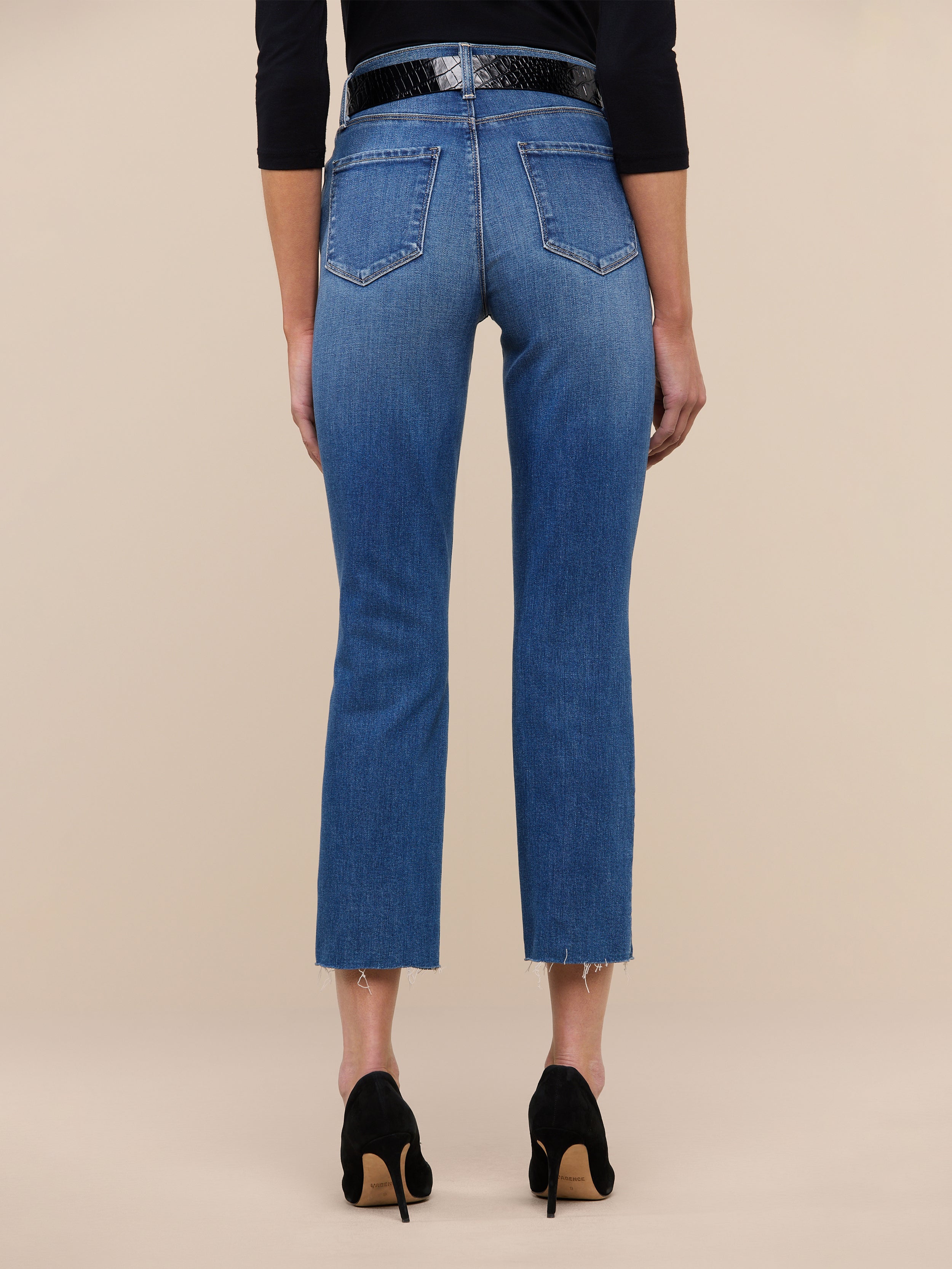 A person stands with their back to the camera against a plain white background, wearing the Sada Slim-Leg Cropped Jean in premium stretch denim with frayed hems, paired with a black belt and black high-heeled shoes.