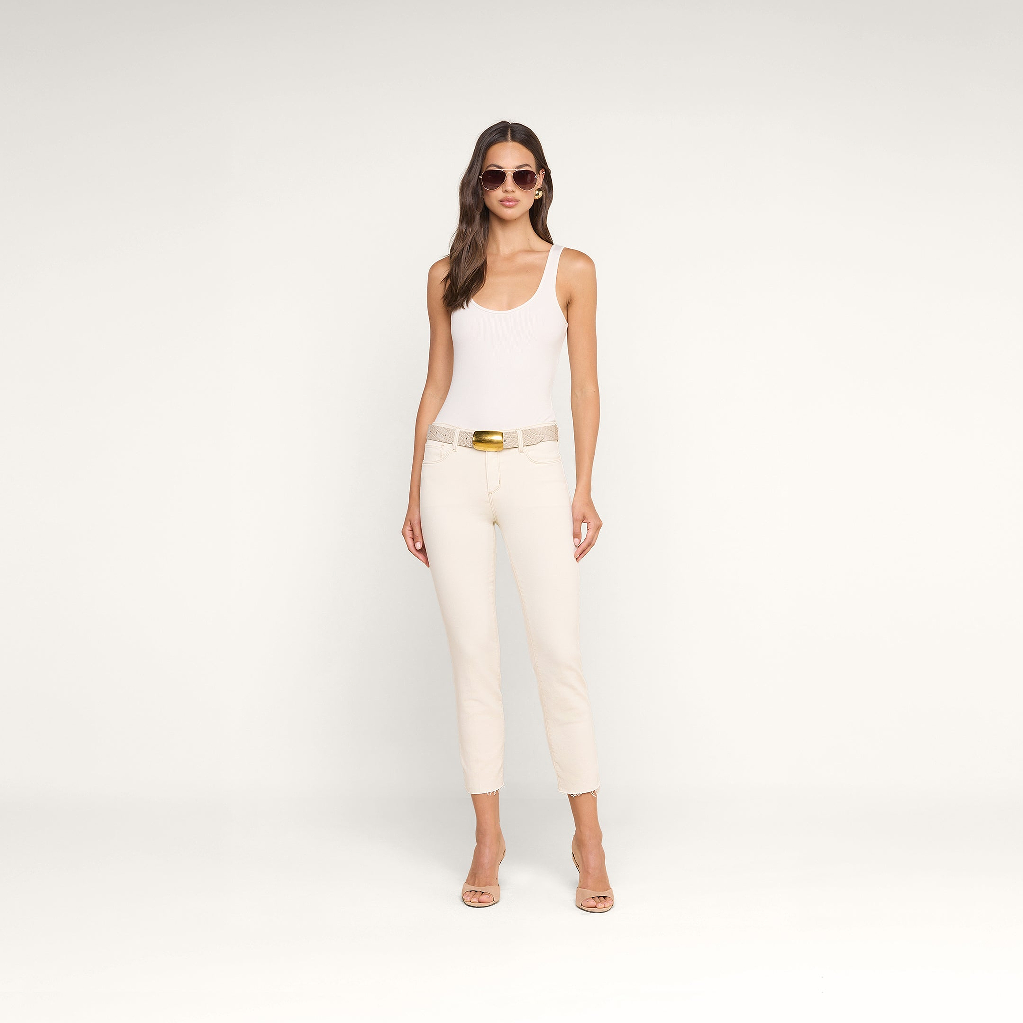 Una donna con occhiali da sole e un top bianco senza maniche indossa i jeans Sada Slim-Leg Cropped, realizzati in denim elasticizzato di alta qualità, abbinati a tacchi color carne su uno sfondo beige.