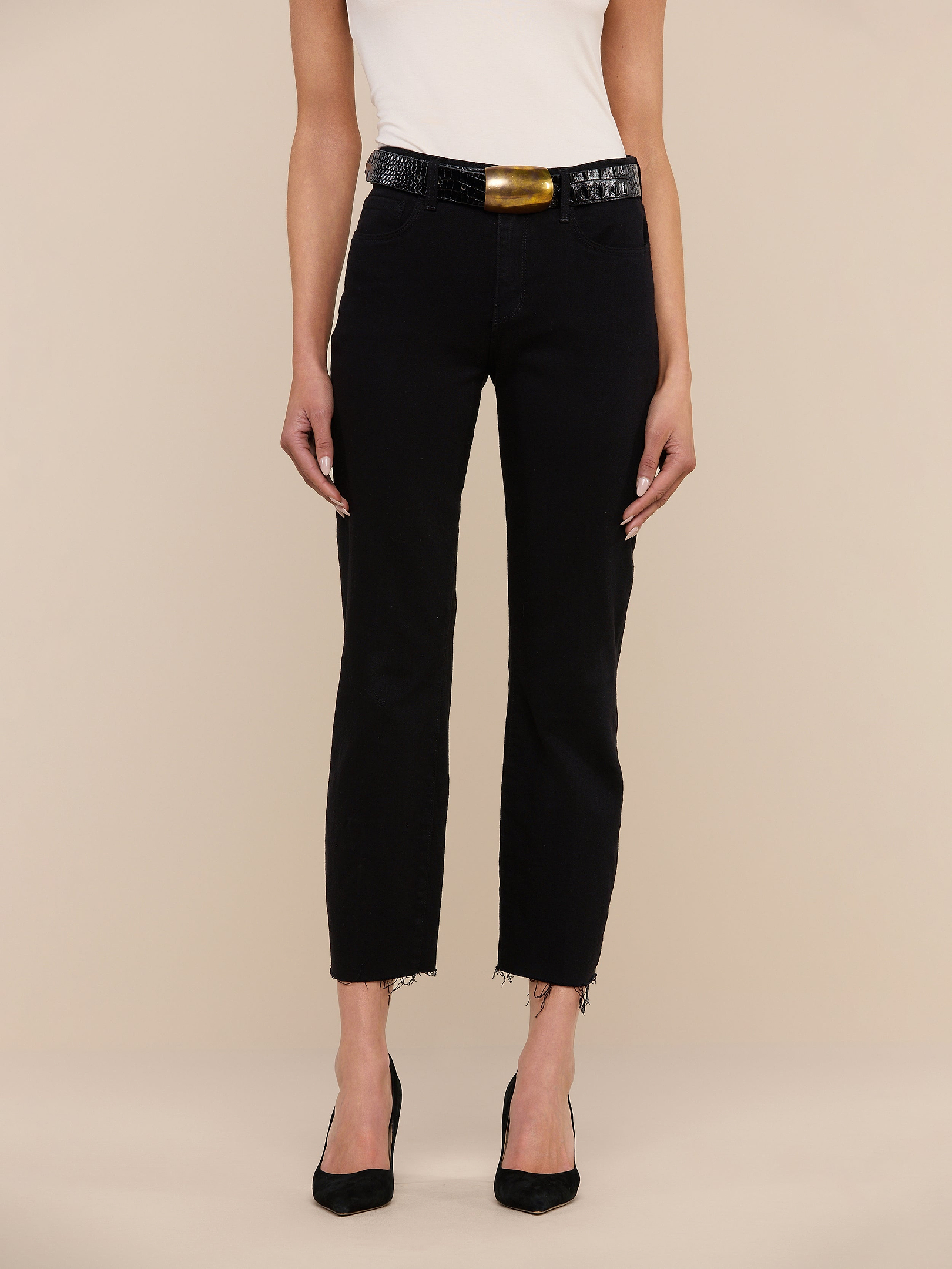 A person stands against a white background, wearing a sleeveless white top and Sada Slim-Leg Cropped Jeans with frayed hems, styled with a black belt featuring a gold buckle and black open-toe heels. Only the body below the shoulders is visible.