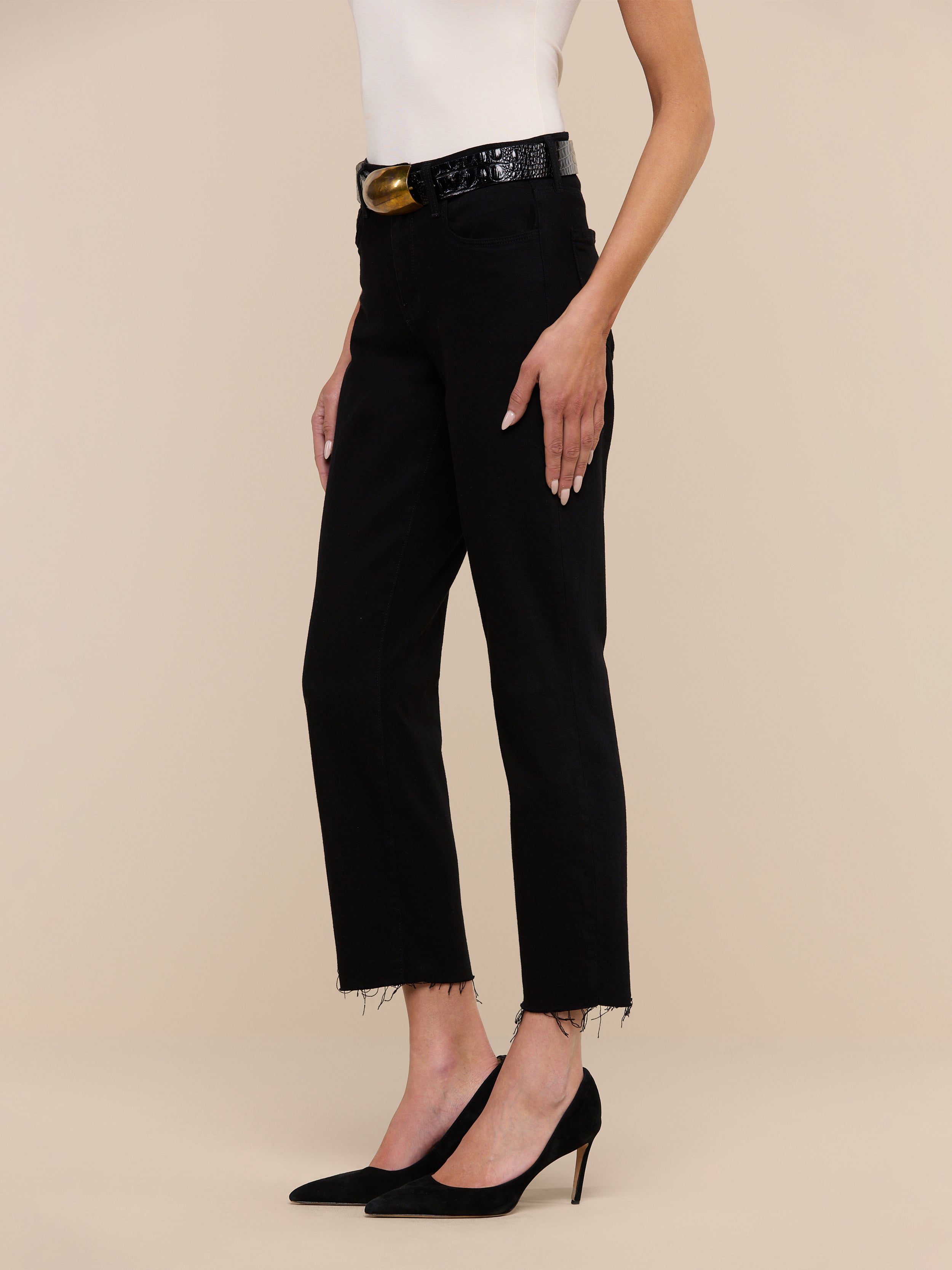A person wearing a sleeveless white top, black Sada Slim-Leg Cropped Jeans with a frayed hem, a black belt, and black pointed-toe heels stands against a plain white background.