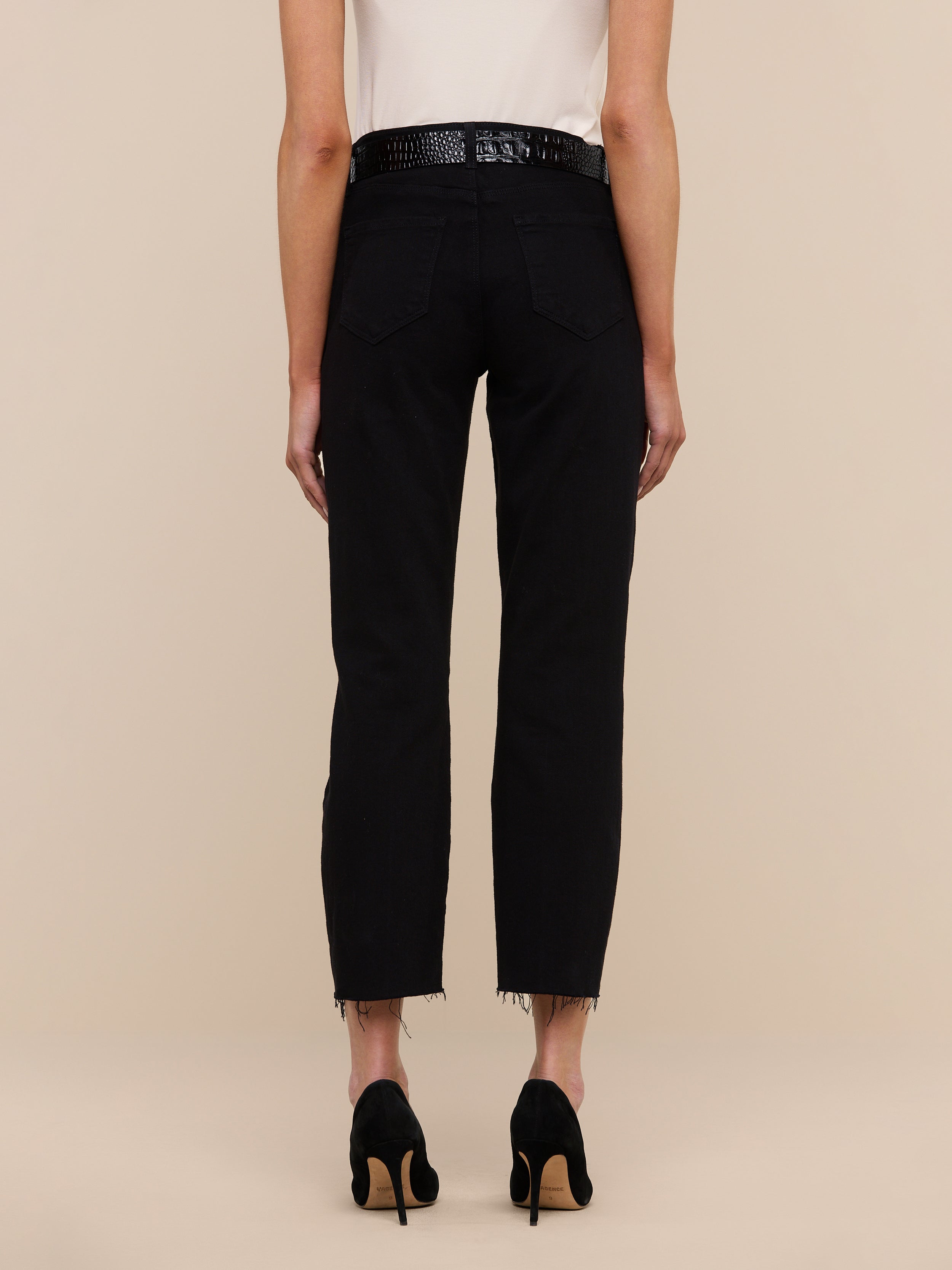 A person stands facing away, wearing the Sada Slim-Leg Cropped Jean—high-rise black jeans with frayed hems—paired with a white sleeveless top and black high-heeled shoes against a plain light background.