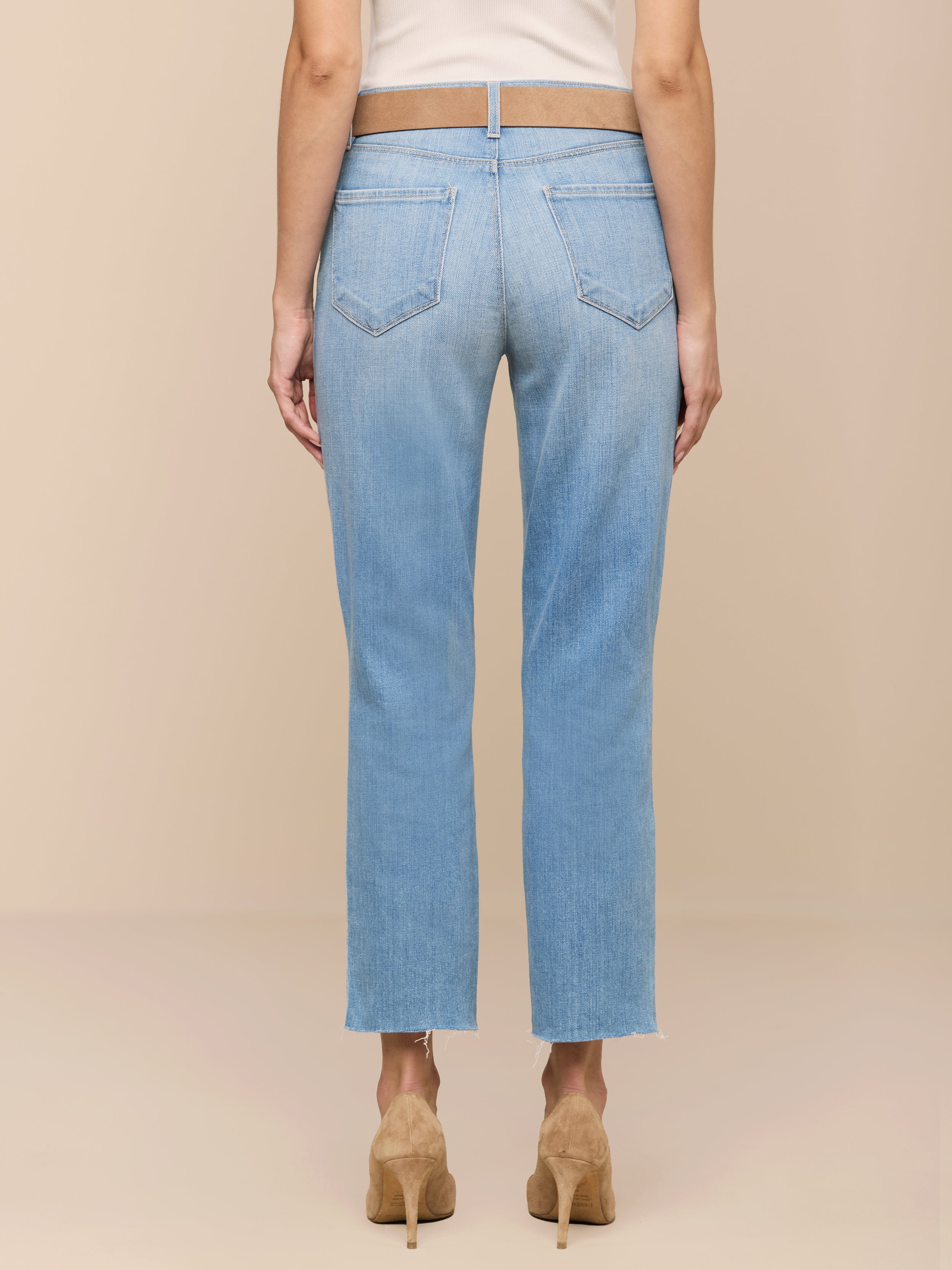 Une personne de dos porte le jean cropped Slim-Leg Sada en délavage clair avec des ourlets bruts et un haut blanc, associé à des chaussures à talons de couleur beige sur un fond beige.