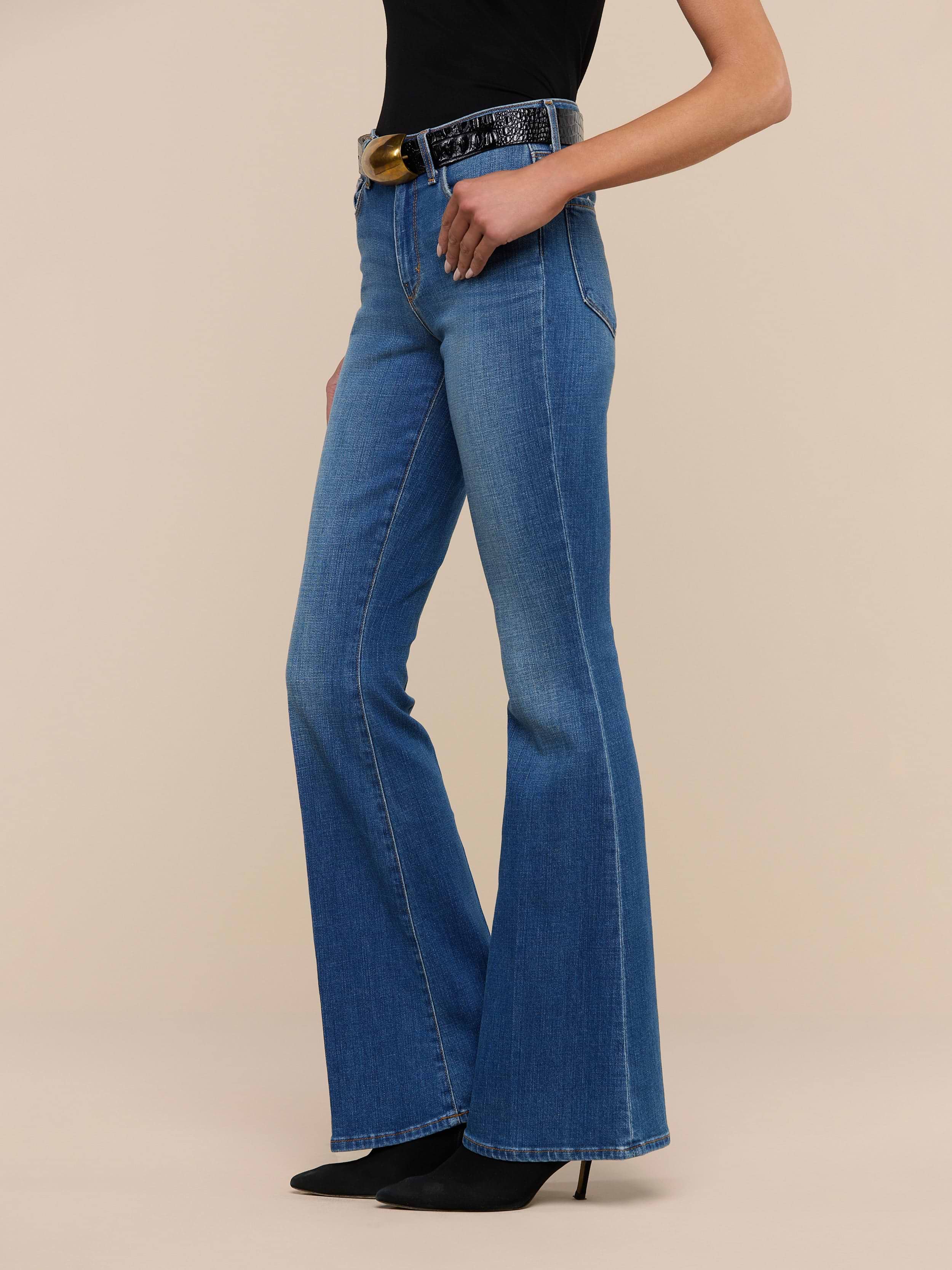 A woman stands against a plain white background wearing the Bell Flare Jean, vintage-inspired blue bell-bottom jeans crafted from premium stretch denim, paired with black pointed-toe heels, a black top, and a black belt featuring a large round gold buckle.