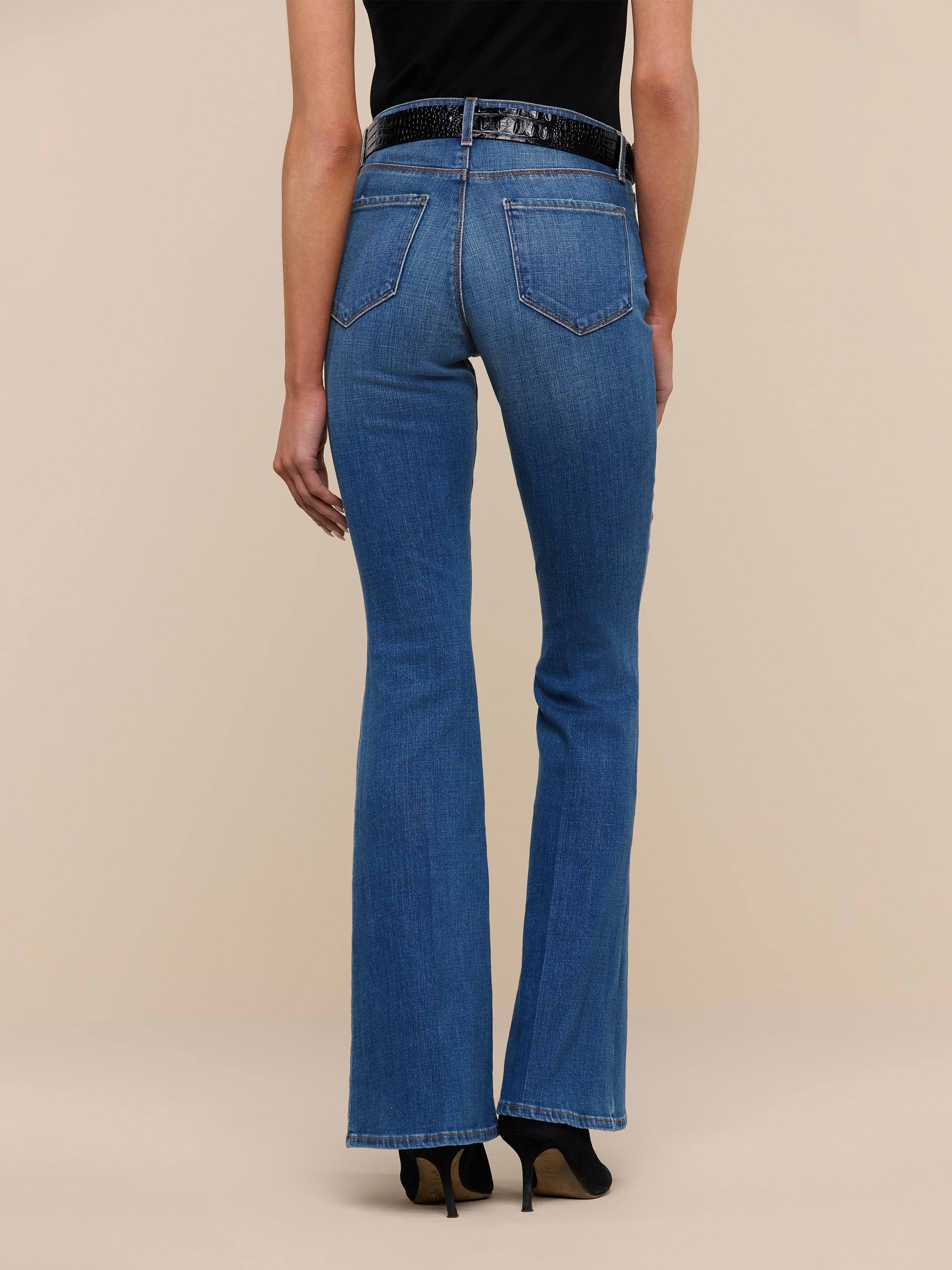 A person stands facing away, wearing the Bell Flare Jean in blue vintage-inspired premium stretch denim, paired with a black top and black heeled shoes against a plain white background.