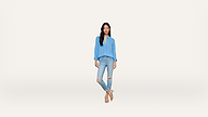 Woman standing against a plain white background, wearing a light blue blouse, ripped light blue jeans, and beige sandals. She has long dark hair and appears relaxed with her hands in her pockets.
