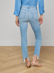 A woman stands with her back to the camera against a plain white background, wearing the High Line Distressed Skinny Jean in a light blue vintage wash with frayed hems, paired with a blue long-sleeve shirt and beige heeled sandals.