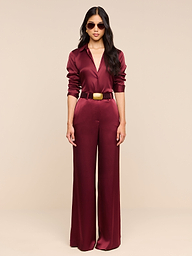 A person wearing the Pilar Silk Wide-Leg Pant in burgundy, paired with a gold belt and sunglasses, stands against a plain white background facing forward with arms relaxed by their sides.
