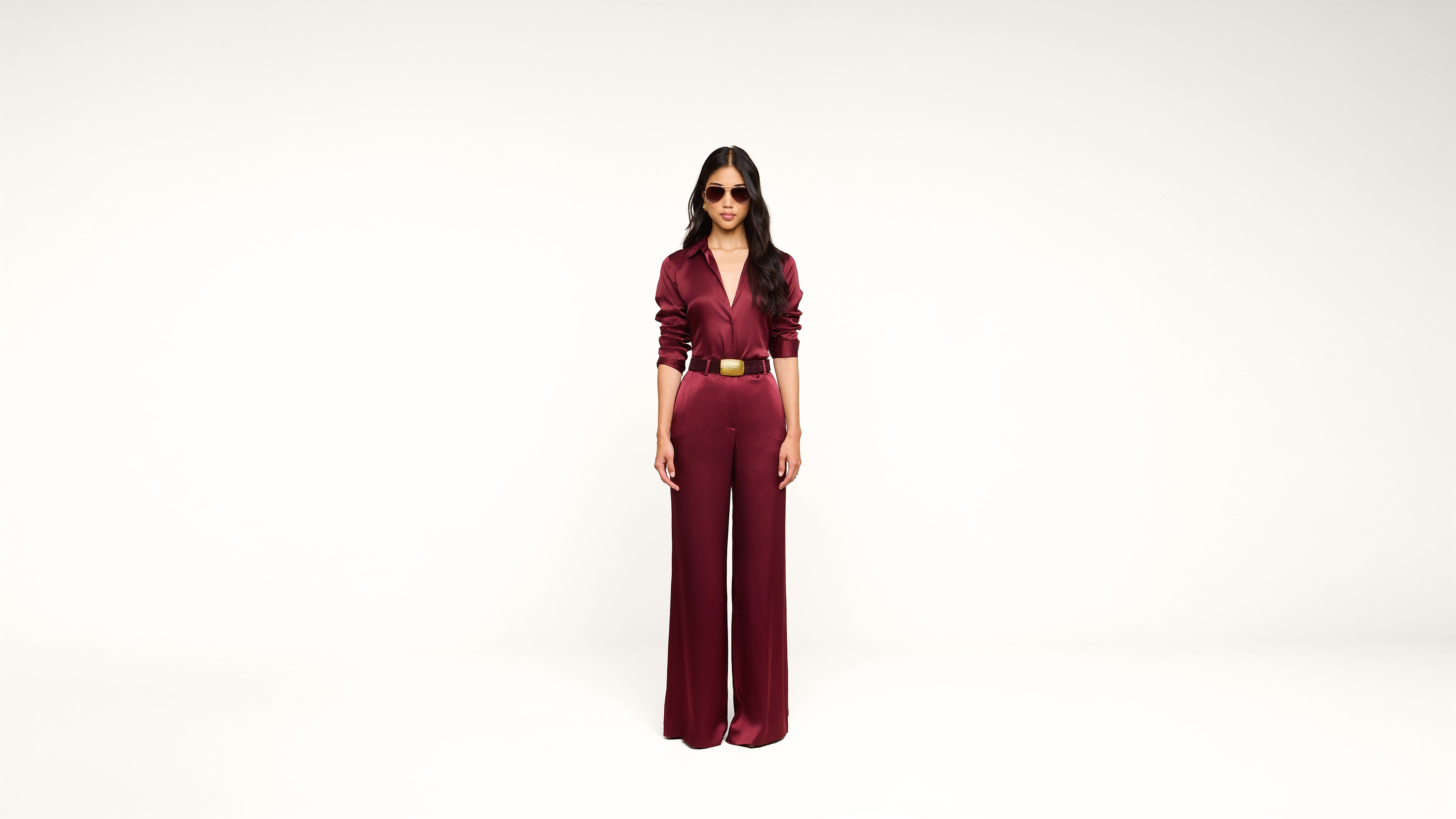 A person wearing sunglasses and a burgundy jumpsuit stands against a plain white background, hands by their sides, facing forward.