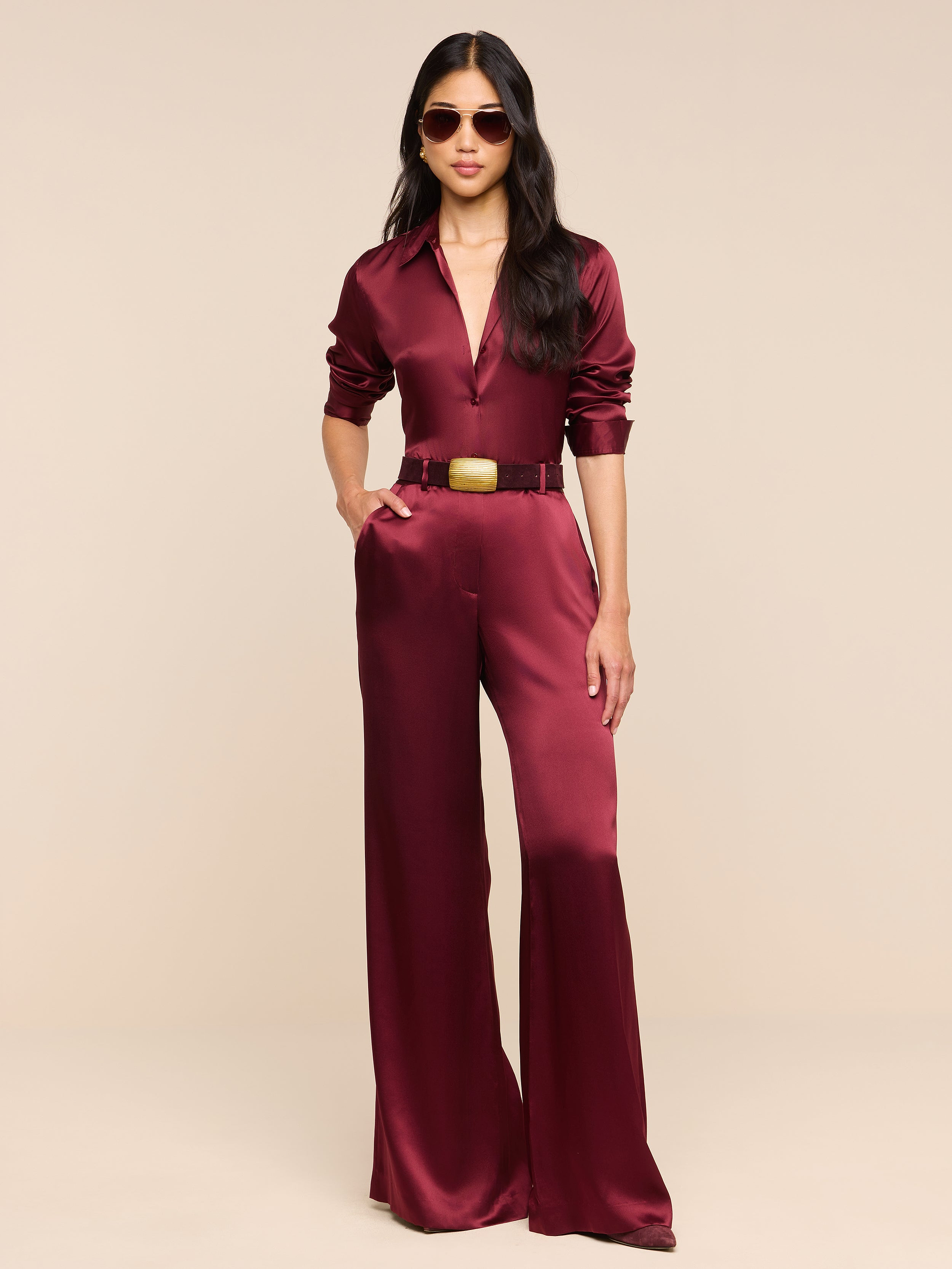 A woman in sunglasses stands confidently against a plain white background, wearing the Pilar Silk Wide-Leg Pant paired with a maroon satin long-sleeve top and a belted waist.
