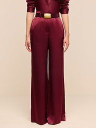 A person wearing the Pilar Silk Wide-Leg Pant, featuring a high-rise, wide-leg silhouette in burgundy satin, paired with a matching long-sleeved top and belt, stands against a plain white background.