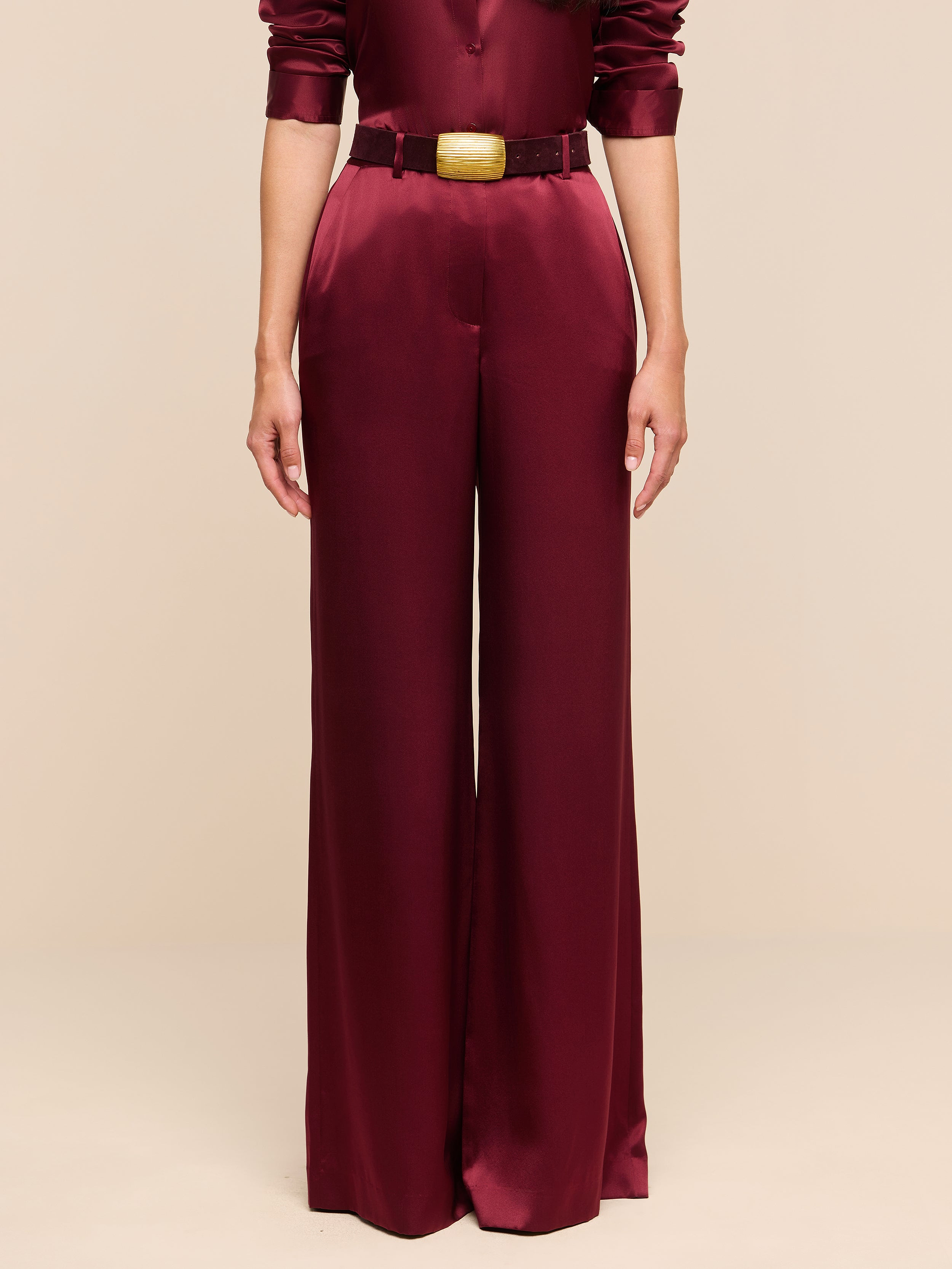 A person wearing the Pilar Silk Wide-Leg Pant, featuring a high-rise, wide-leg silhouette in burgundy satin, paired with a matching long-sleeved top and belt, stands against a plain white background.