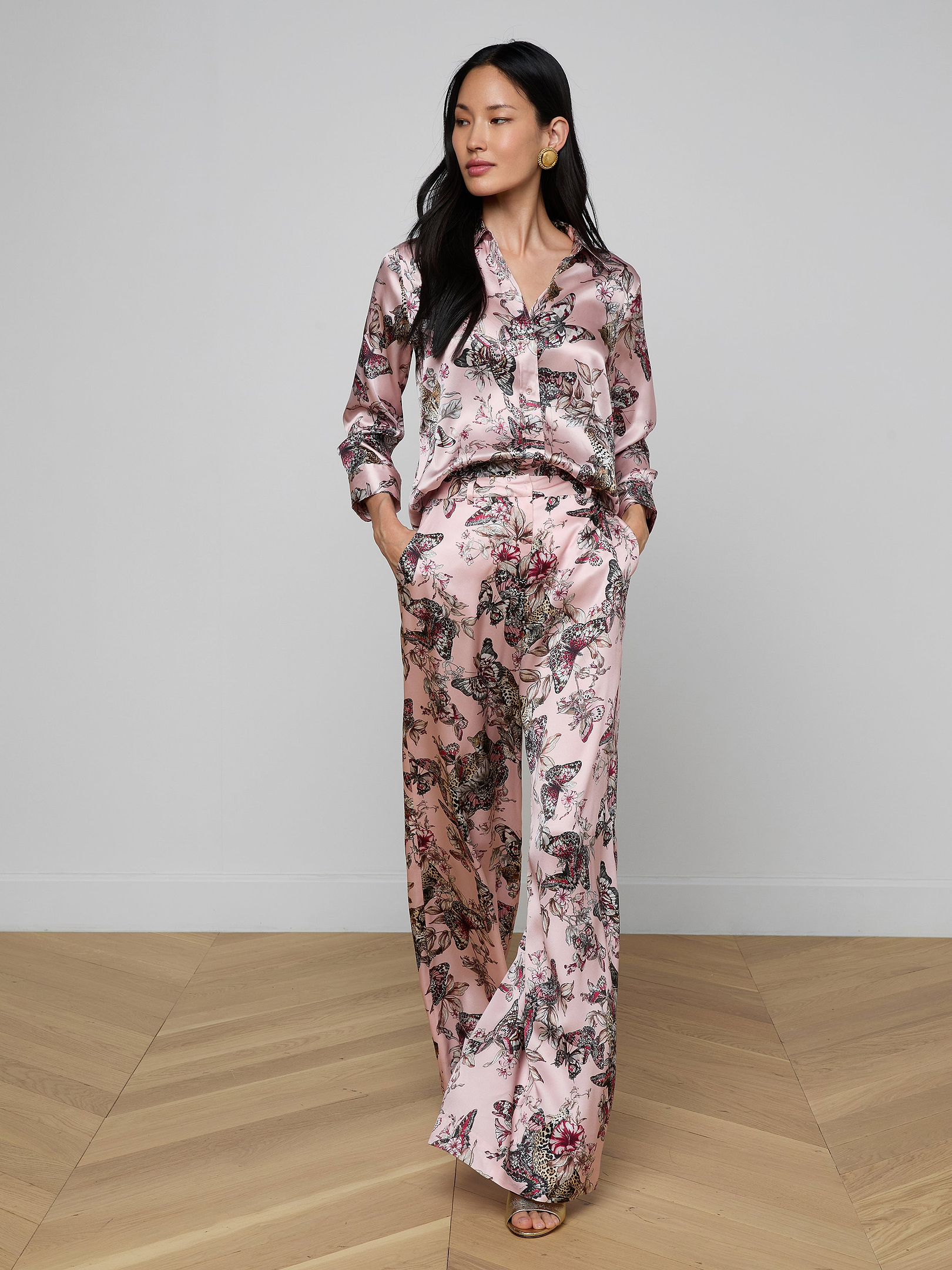 A woman wearing the Pilar Silk Wide-Leg Pant, featuring a silky, light pink fabric with floral patterns and a high-rise fit, stands with her hands in her pockets against a plain off-white background.