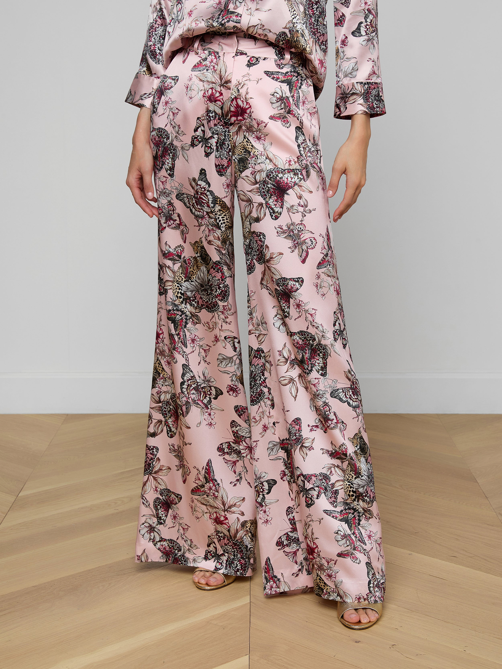 Wearing the Pilar Silk Wide-Leg Pant, styled with a coordinating long-sleeve top in pink fabric featuring a vibrant butterfly jungle print, this look is completed with open-toe shoes. The image is cropped to show only from the shoulders down.