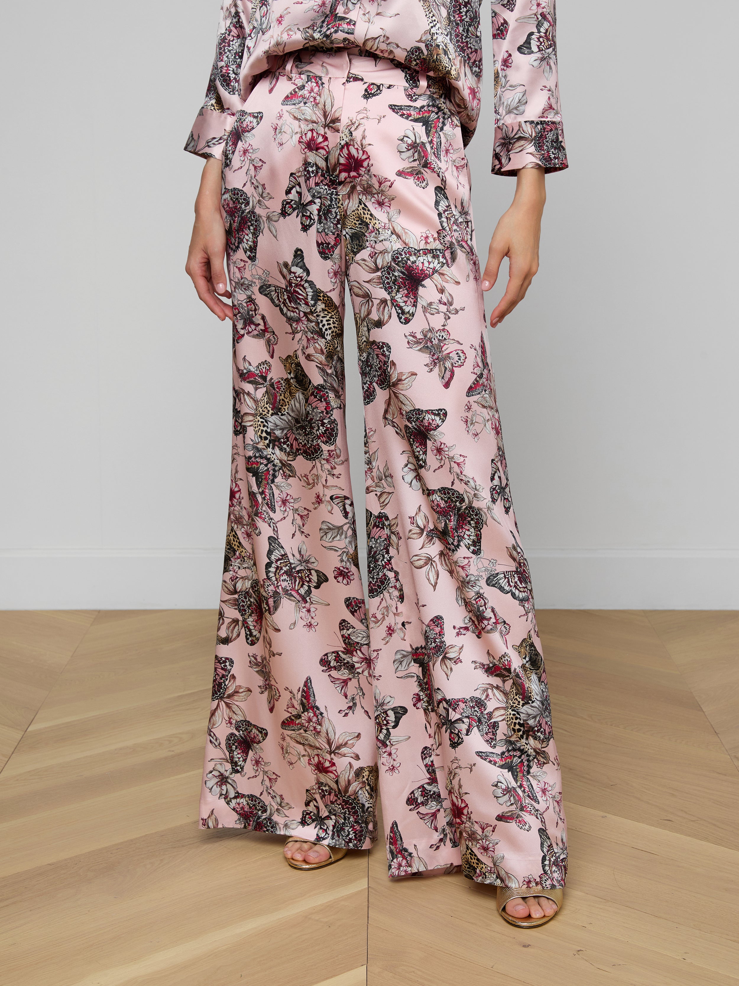 Wearing the Pilar Silk Wide-Leg Pant, styled with a coordinating long-sleeve top in pink fabric featuring a vibrant butterfly jungle print, this look is completed with open-toe shoes. The image is cropped to show only from the shoulders down.