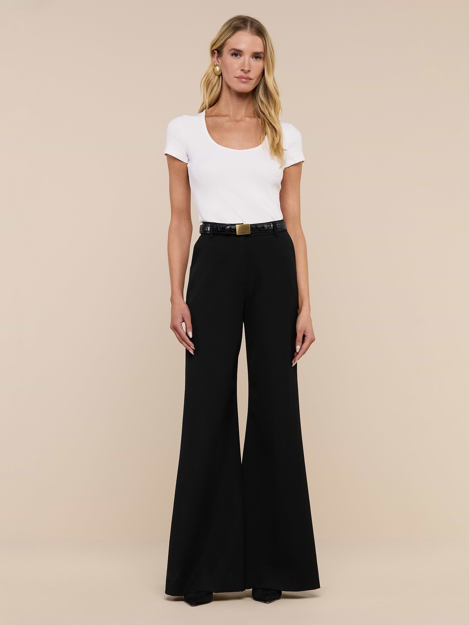 A woman stands against a plain white background, wearing a white short-sleeve top tucked into the Pilar Wide-Leg Pant, crafted from four-way stretch suiting and finished with a black belt and black shoes.
