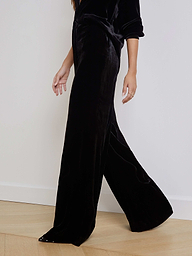 A person shown from the shoulders down walks against a plain white background, wearing the black Pilar Velvet Wide-Leg Pant with a matching top.