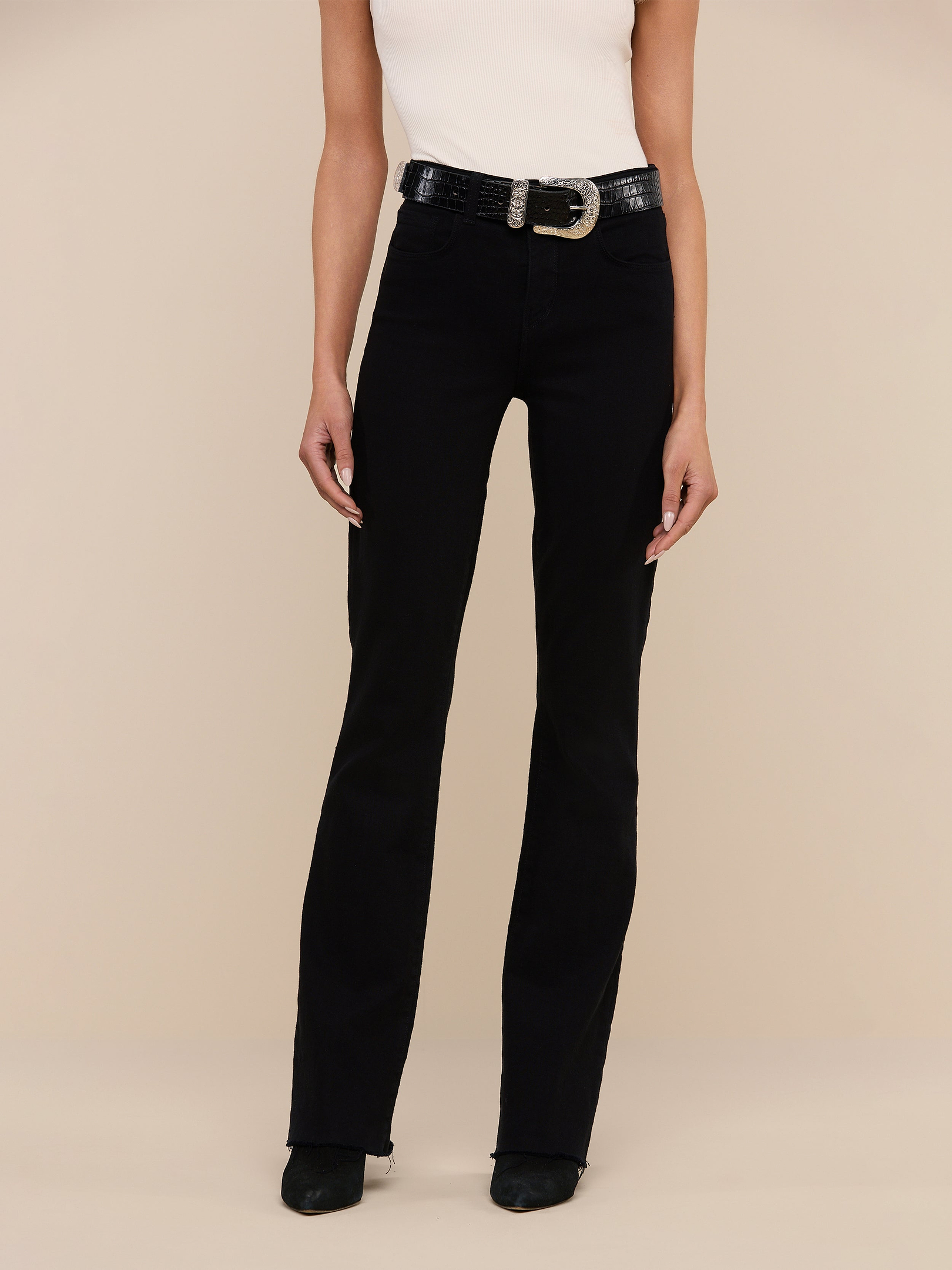A person wearing the Ruth Straight-Leg Jean in black stretch denim, styled with a white sleeveless top, black shoes, and a black belt with a large buckle stands against a plain white background.