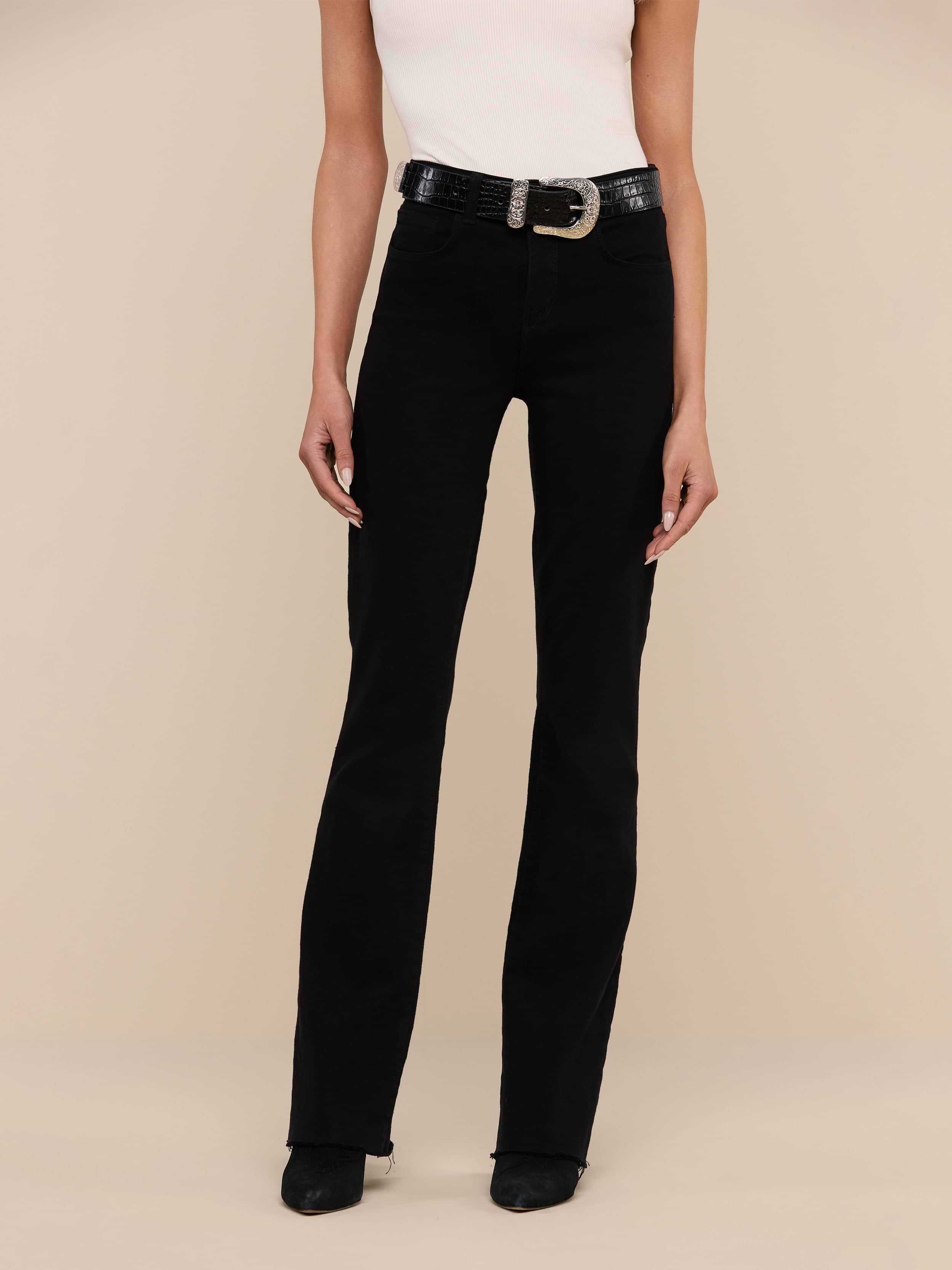 A person wearing the Ruth Straight-Leg Jean in black stretch denim, styled with a white sleeveless top, black shoes, and a black belt with a large buckle stands against a plain white background.