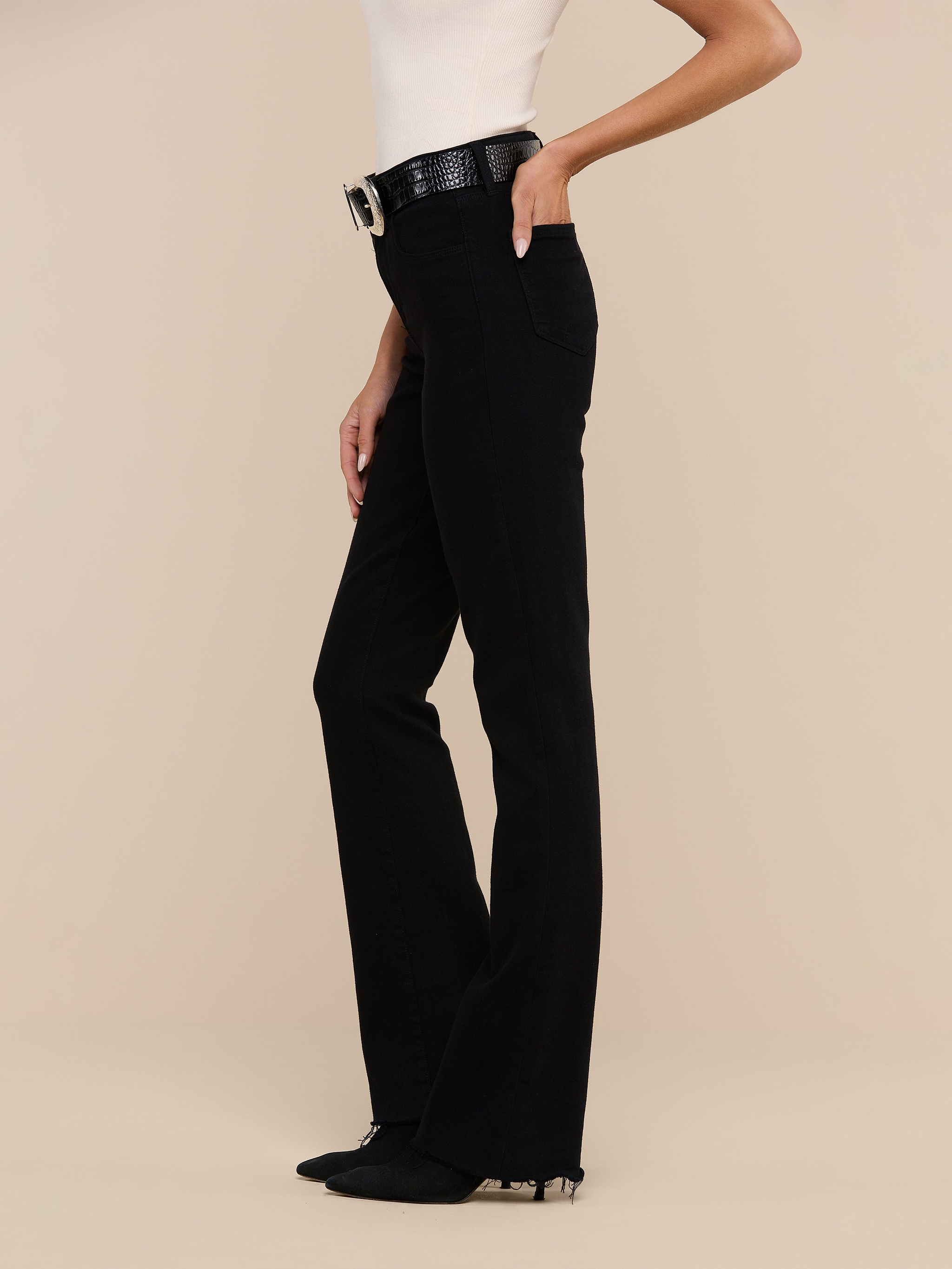 Woman wearing a sleeveless top, Ruth Straight-Leg Jean in black high-rise stretch denim, a black belt with a large buckle, and black shoes, standing against a plain white background.
