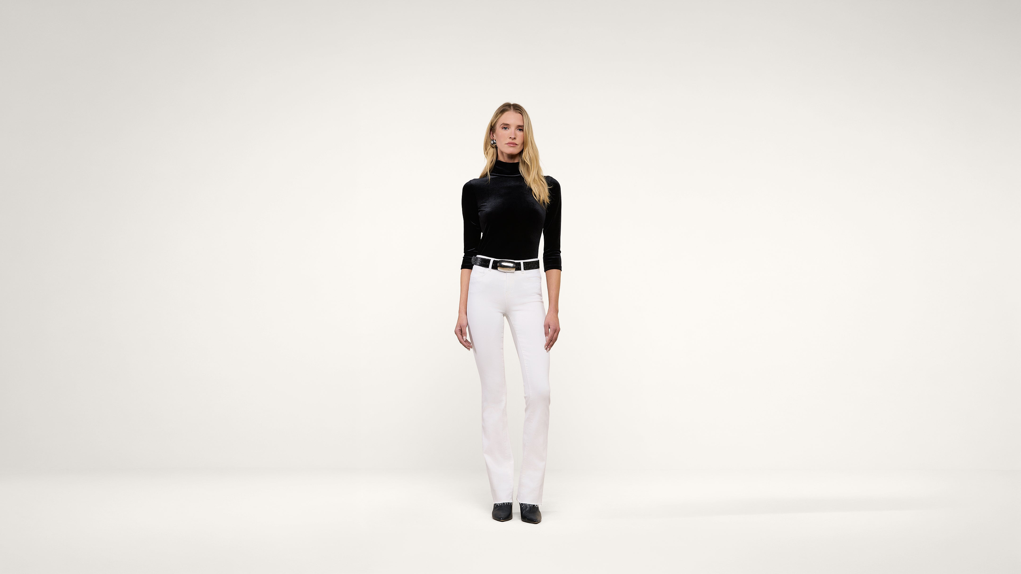 A woman with long blonde hair stands facing forward, wearing a black turtleneck top, white pants, black belt, and black shoes, against a plain white background.