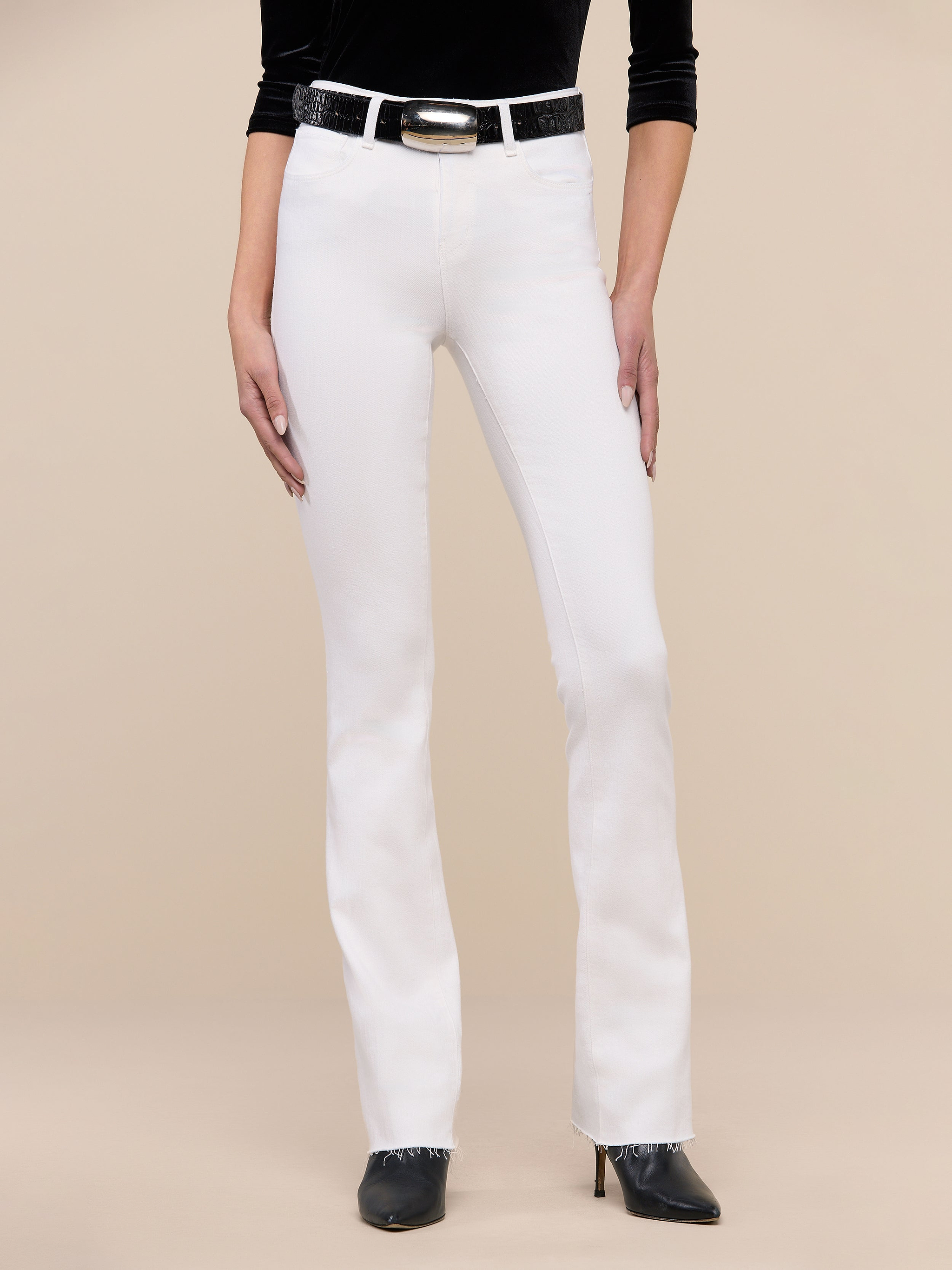 A person wearing the Ruth Straight-Leg Jean in white stretch denim, paired with a black top, a black belt with a silver buckle, and black pointed-toe heels stands against a plain white background.