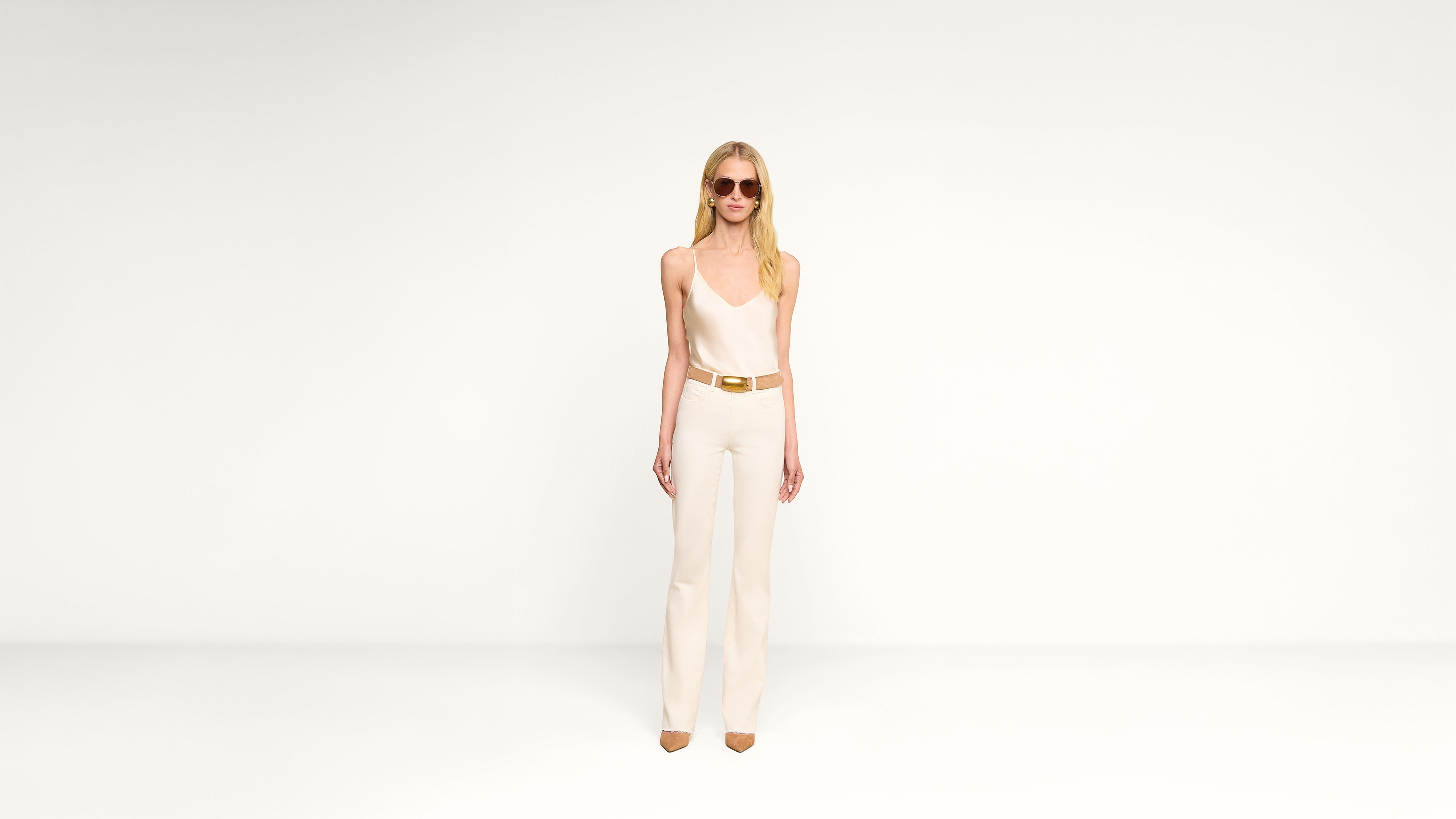 A woman wearing sunglasses, a white sleeveless top, white pants, a gold belt, and tan shoes stands against a plain white background.
