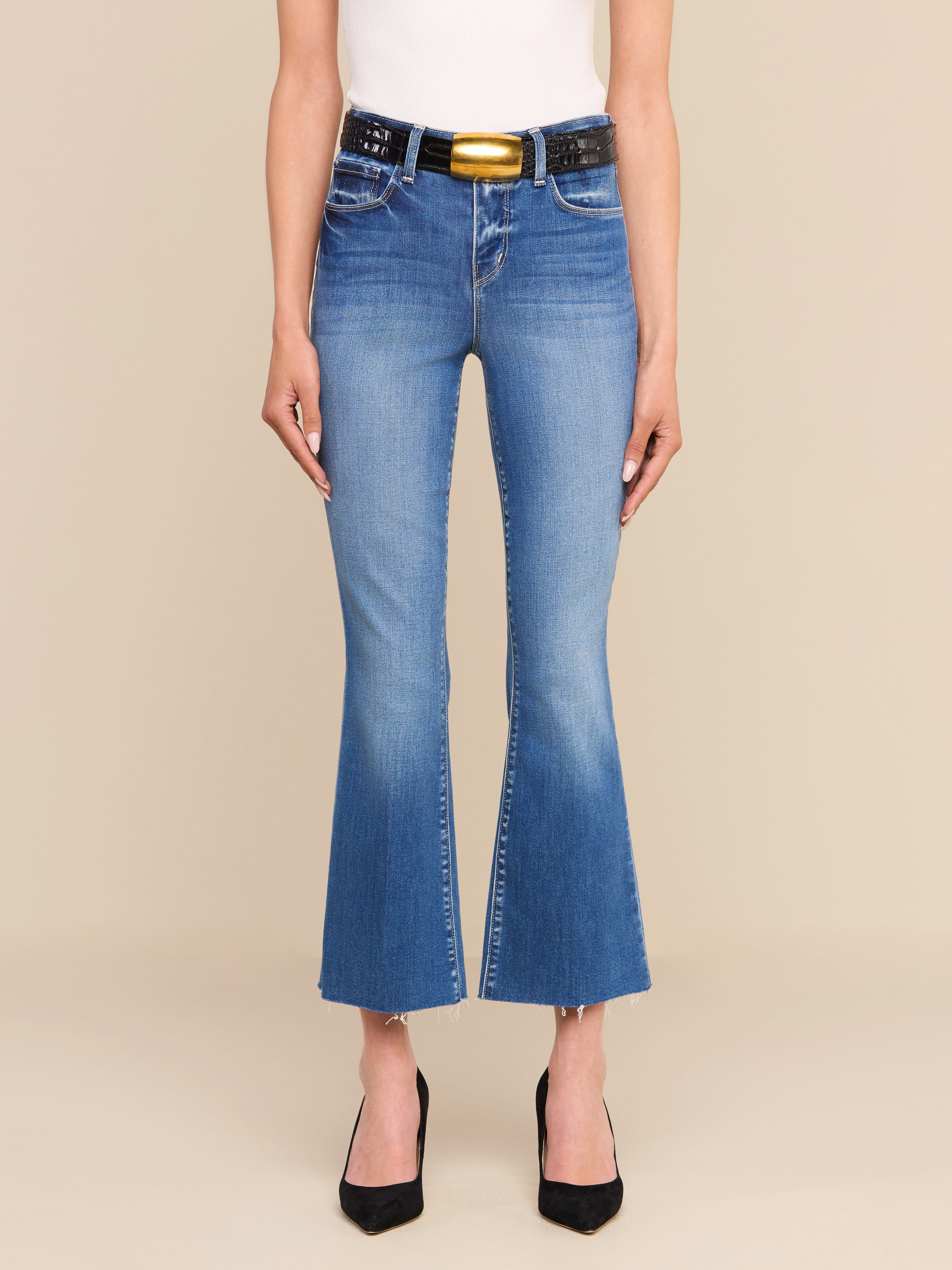 Una persona che indossa i jeans Kendra Cropped Flare — un modello blu a zampa d’elefante dal taglio corto con orlo grezzo, realizzati in denim elasticizzato di alta qualità — abbinati a una cintura nera con una grande fibbia dorata, un top bianco senza maniche e tacchi neri a punta, in piedi su uno sfondo beige.