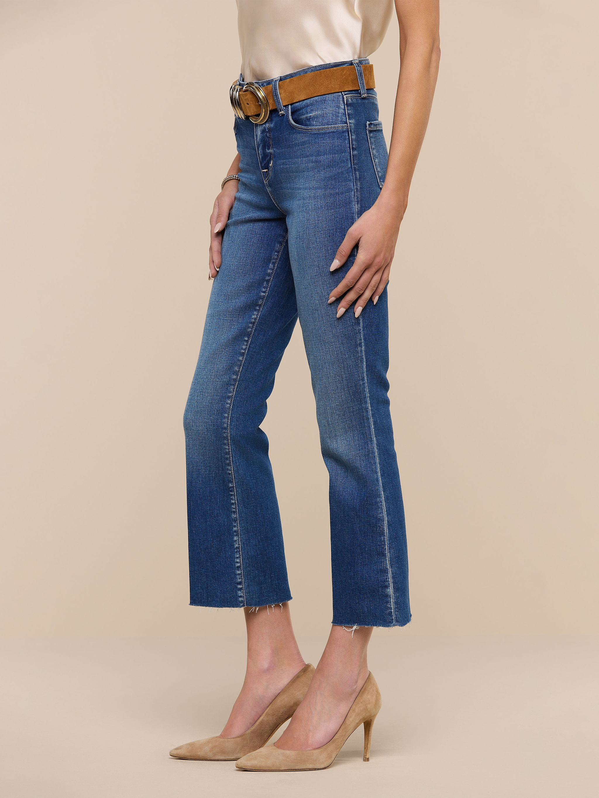 A person wearing the Kendra Cropped Flare Jean in blue premium stretch denim, paired with a black belt featuring a gold buckle, a white top, and black high-heeled shoes, standing against a plain white background.