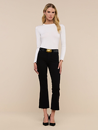 A woman stands facing forward, wearing a white long-sleeve top and the Kendra Cropped Flare Jean—premium high-rise cropped flare jeans in a black hue with frayed hems—paired with a black belt featuring a gold buckle and black heels, against a plain white background.