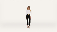 A woman stands facing forward against a plain white background, wearing a white long-sleeve top, black pants with a gold-buckled belt, and black heels.