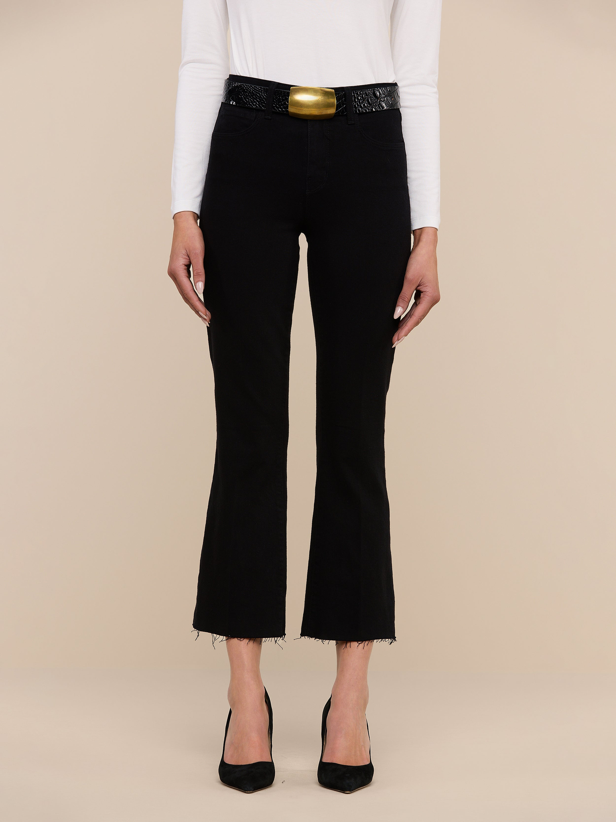 A person wearing a white long-sleeve top, the Kendra Cropped Flare Jean in a rich black wash with frayed hems, a wide black belt featuring a large gold buckle, and black heels stands against a plain white background.