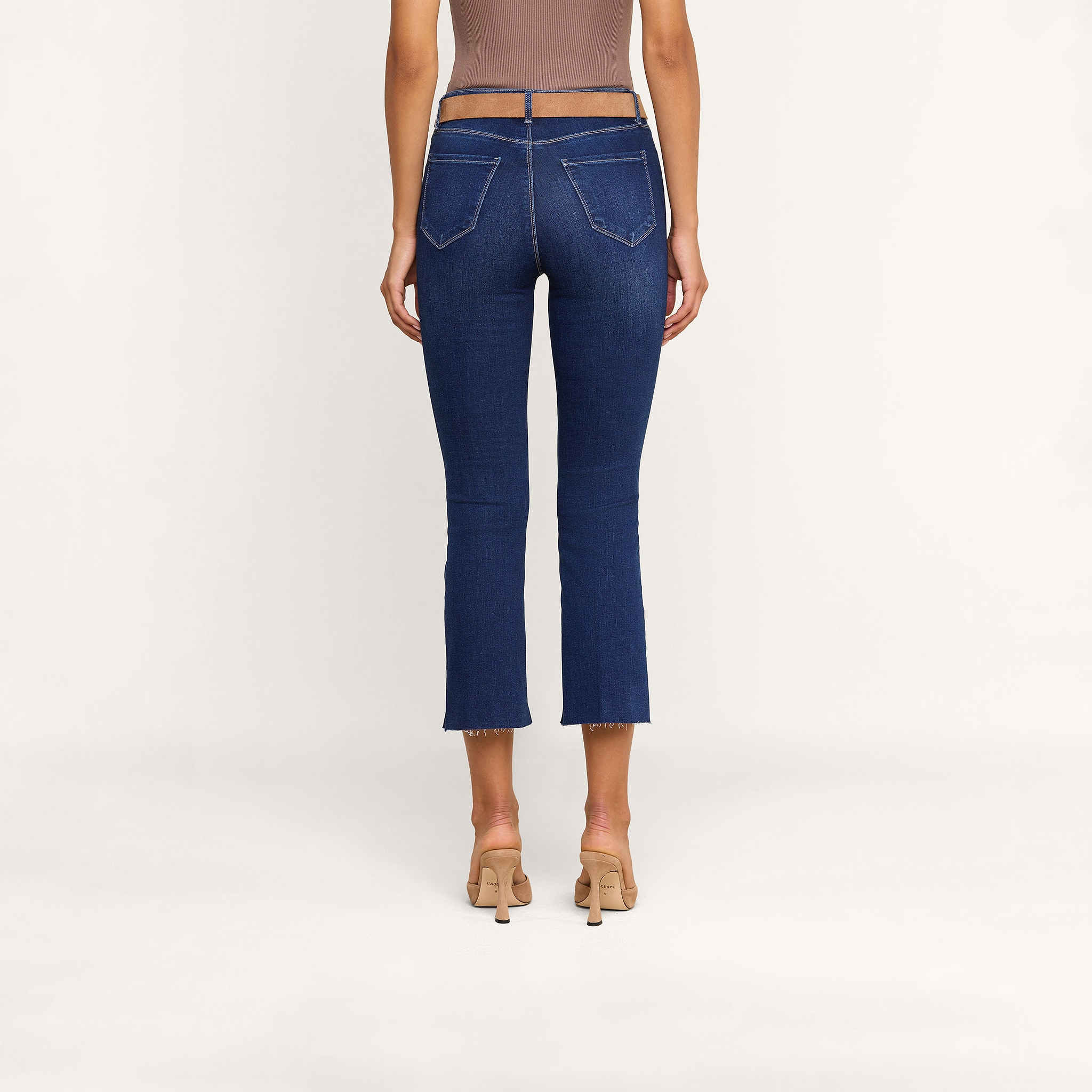 Una donna è di spalle e indossa un paio di jeans Kendra Cropped Flare in denim elasticizzato di alta qualità con orli sfrangiati e tasche posteriori, un top senza maniche color taupe e sandali con tacco color carne, su uno sfondo beige.