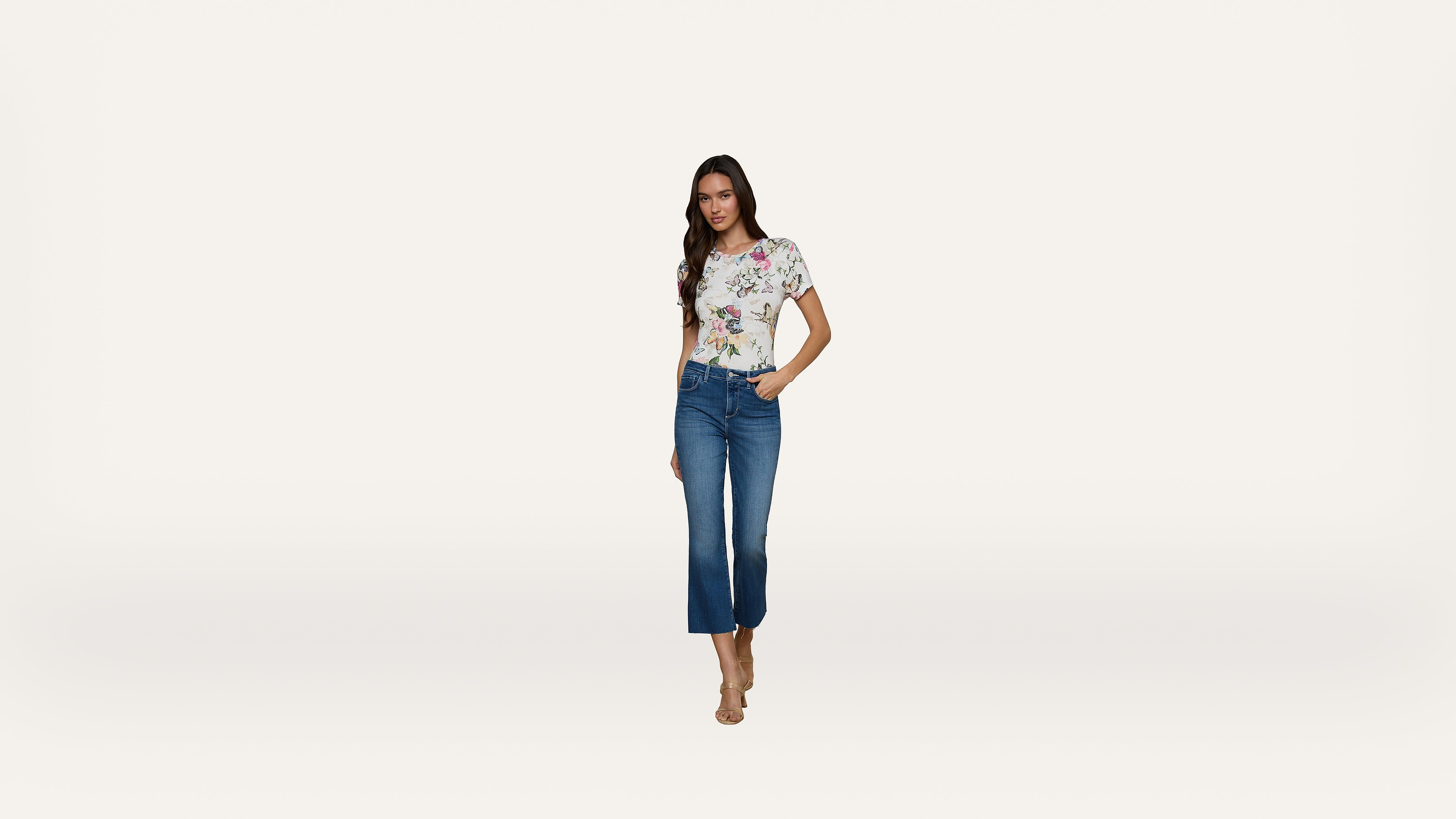 A woman stands against a plain white background, wearing a floral short-sleeve top, blue cropped jeans, and nude heeled sandals.