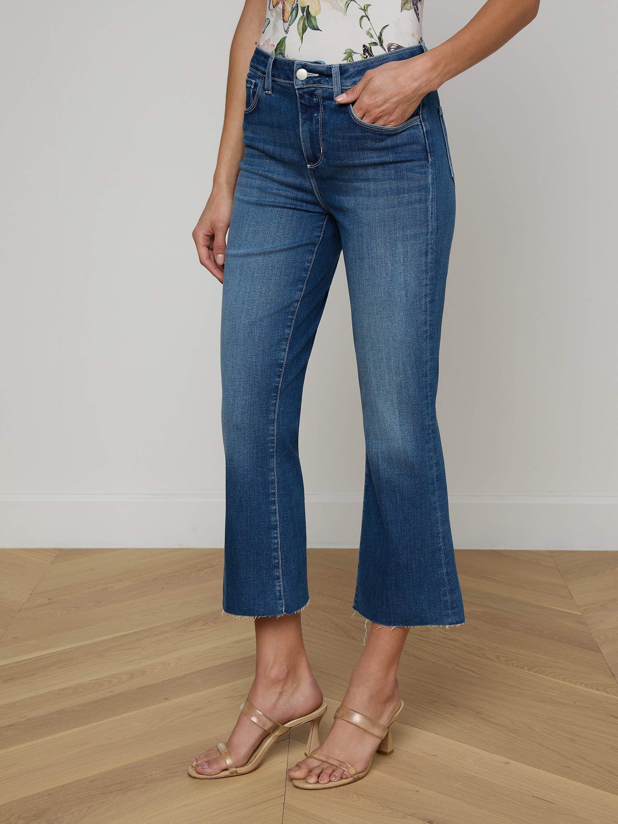 A person models the Kendra Cropped Flare Jean in a dark denim wash, paired with a floral top and beige heeled sandals, posing against a plain white background with only the lower half of the body visible.