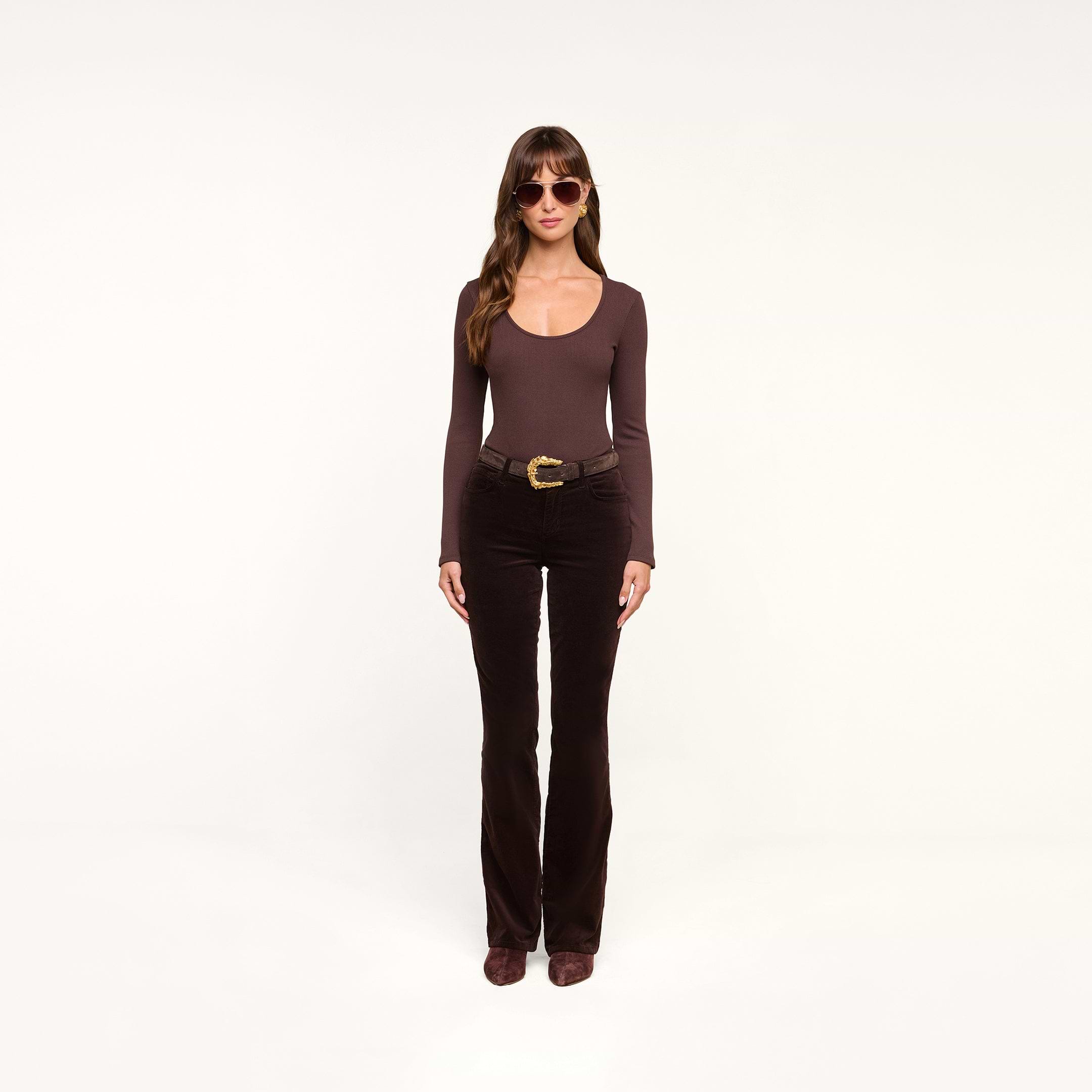 A woman wearing sunglasses, a long-sleeve dark java brown stretch velvet top, the Stevie Velvet Jean in brown with a belt, and brown shoes stands facing forward against a plain white background.