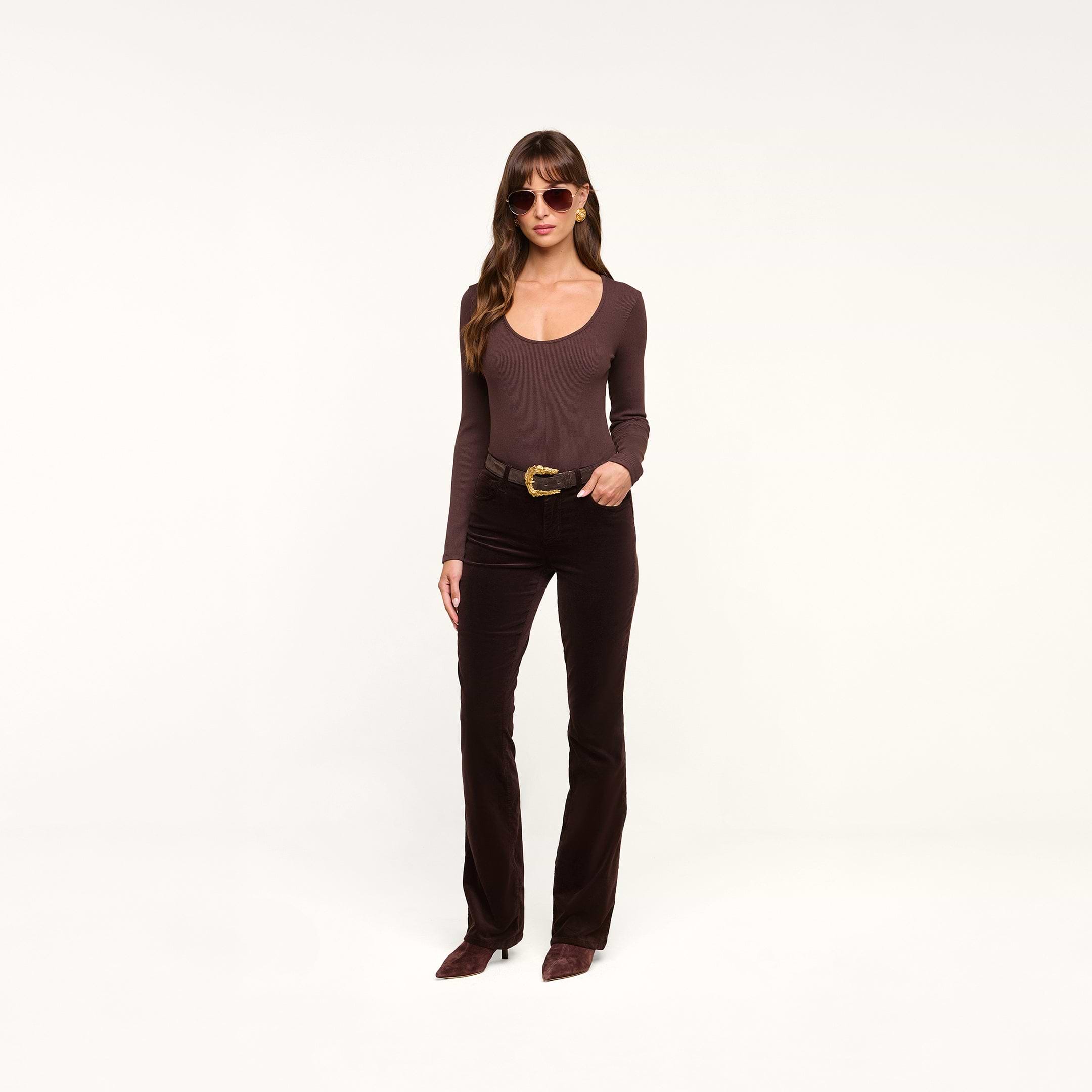 A woman wearing sunglasses, a fitted brown long-sleeve top, the Stevie Velvet Jean in slim straight leg dark java brown stretch velvet, and brown shoes stands against a plain white background.