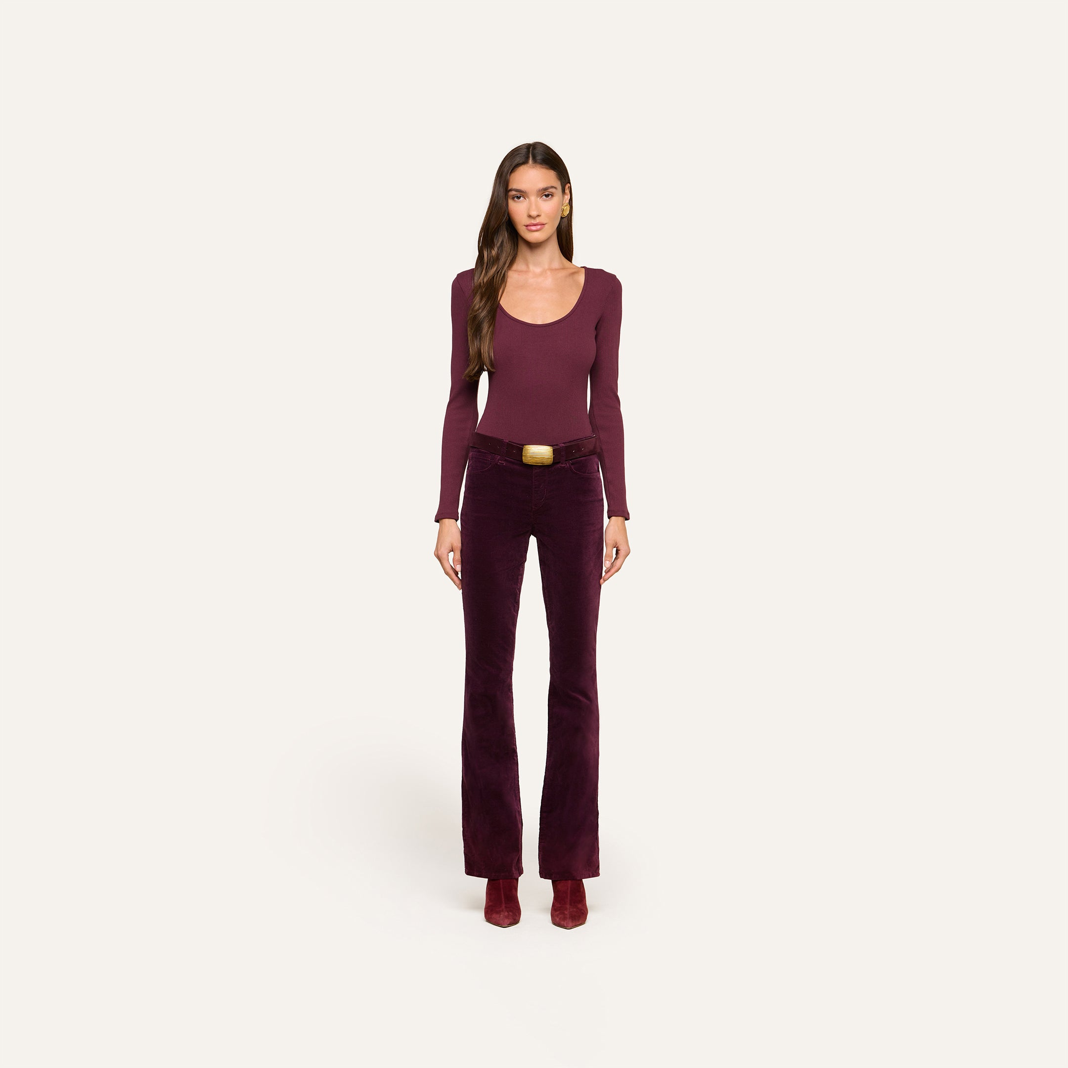 A woman stands facing forward against a plain off-white background, wearing a long-sleeve burgundy top, heeled boots, and high-rise Stevie Velvet Jeans with a belt.