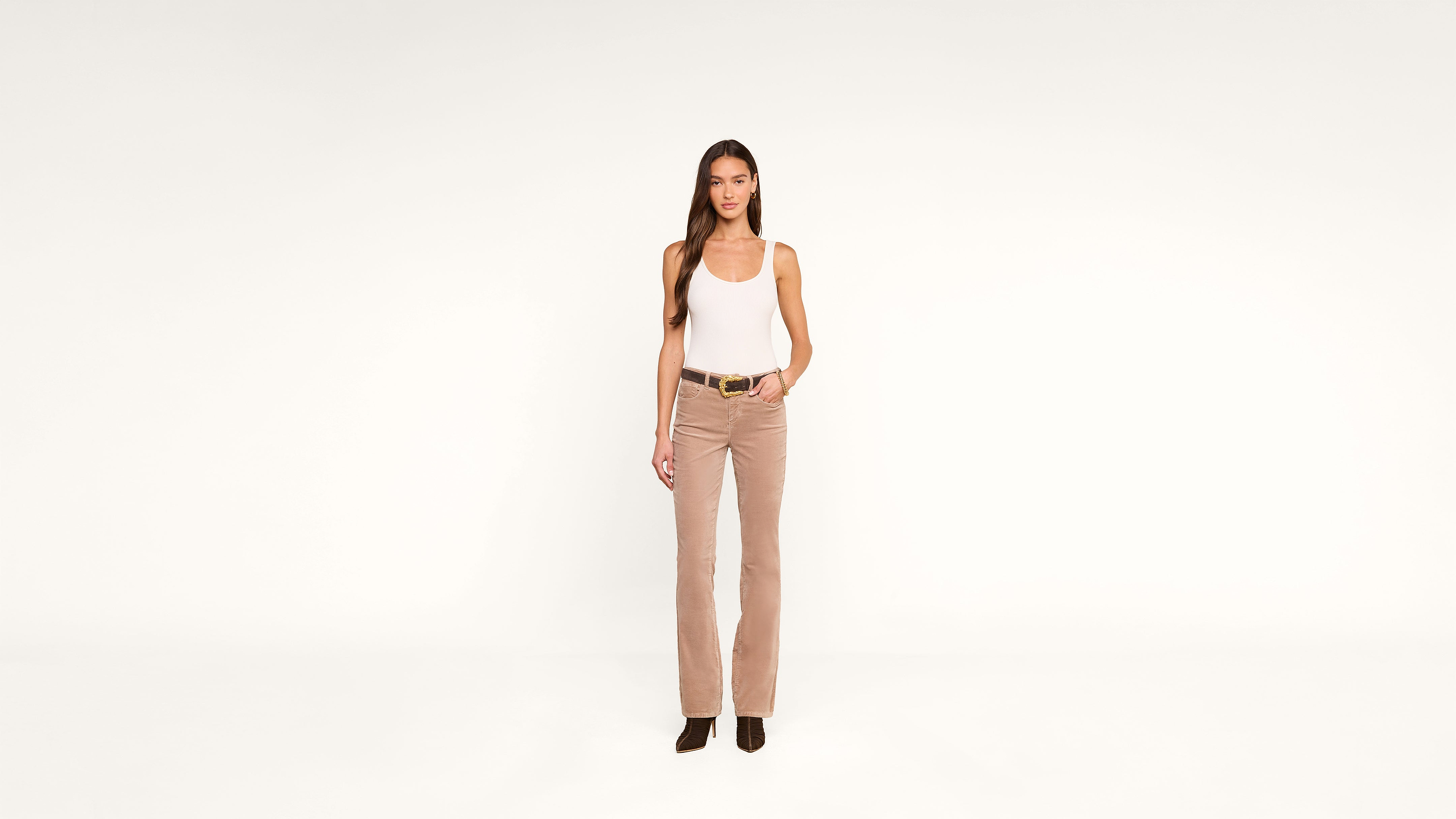 Woman stands against a plain white background, wearing a white tank top, beige corduroy pants, black belt, and black shoes, with one hand in her pocket.